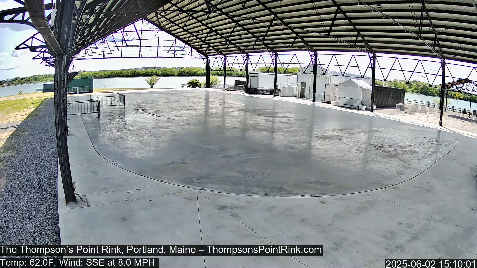 An empty, paved rink sits under a metal-framed awning overlooking a calm body of water on a sunny day.