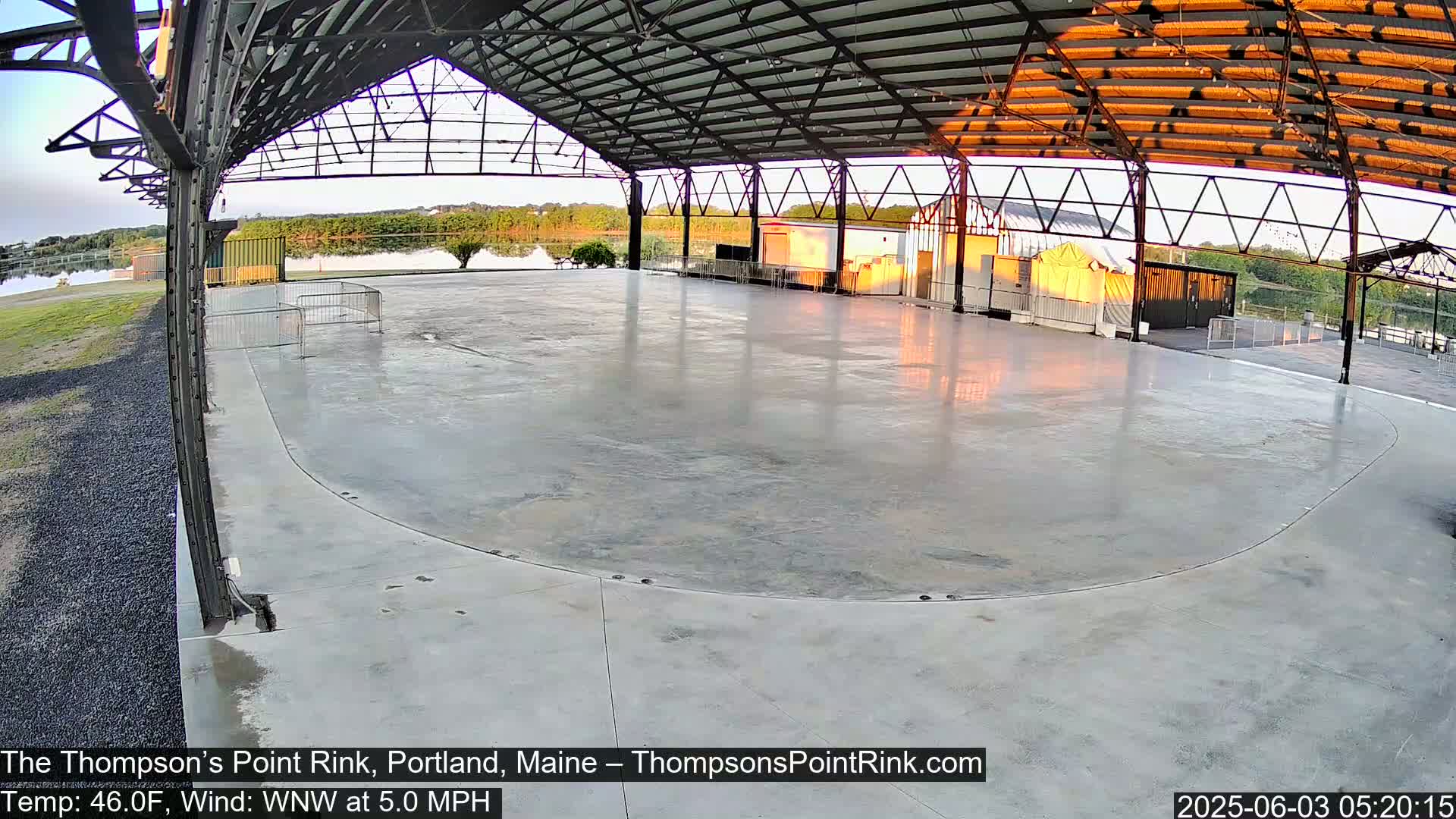 A large, empty concrete rink sits under a metal-framed structure, overlooking a calm body of water at sunrise.