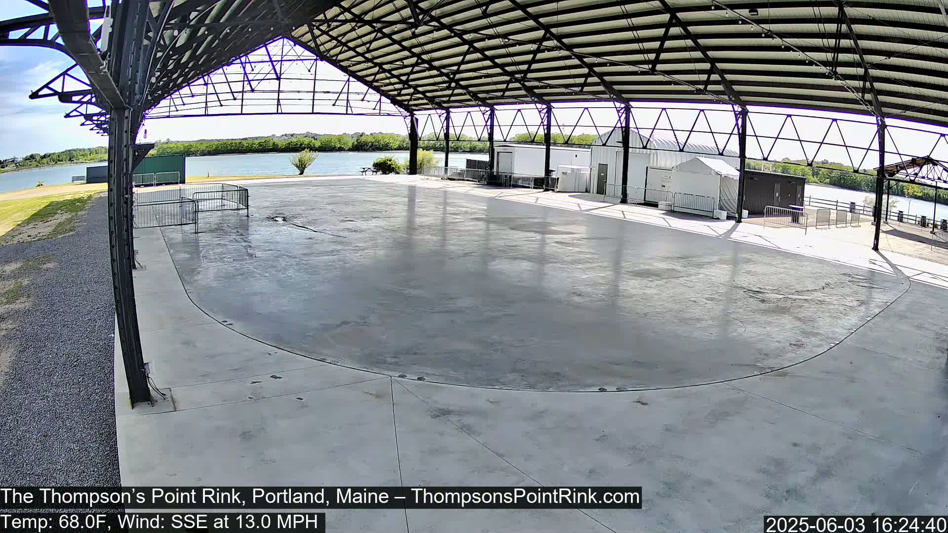 An empty, paved rink sits under a metal-framed open-air shelter on a sunny day with a calm river visible in the background.