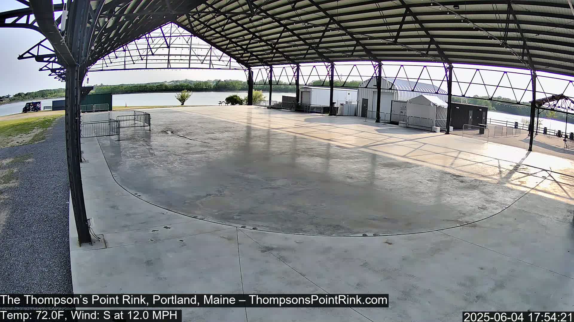 An empty, paved outdoor rink sits under a metal shelter overlooking a calm body of water on a sunny day with a south wind.