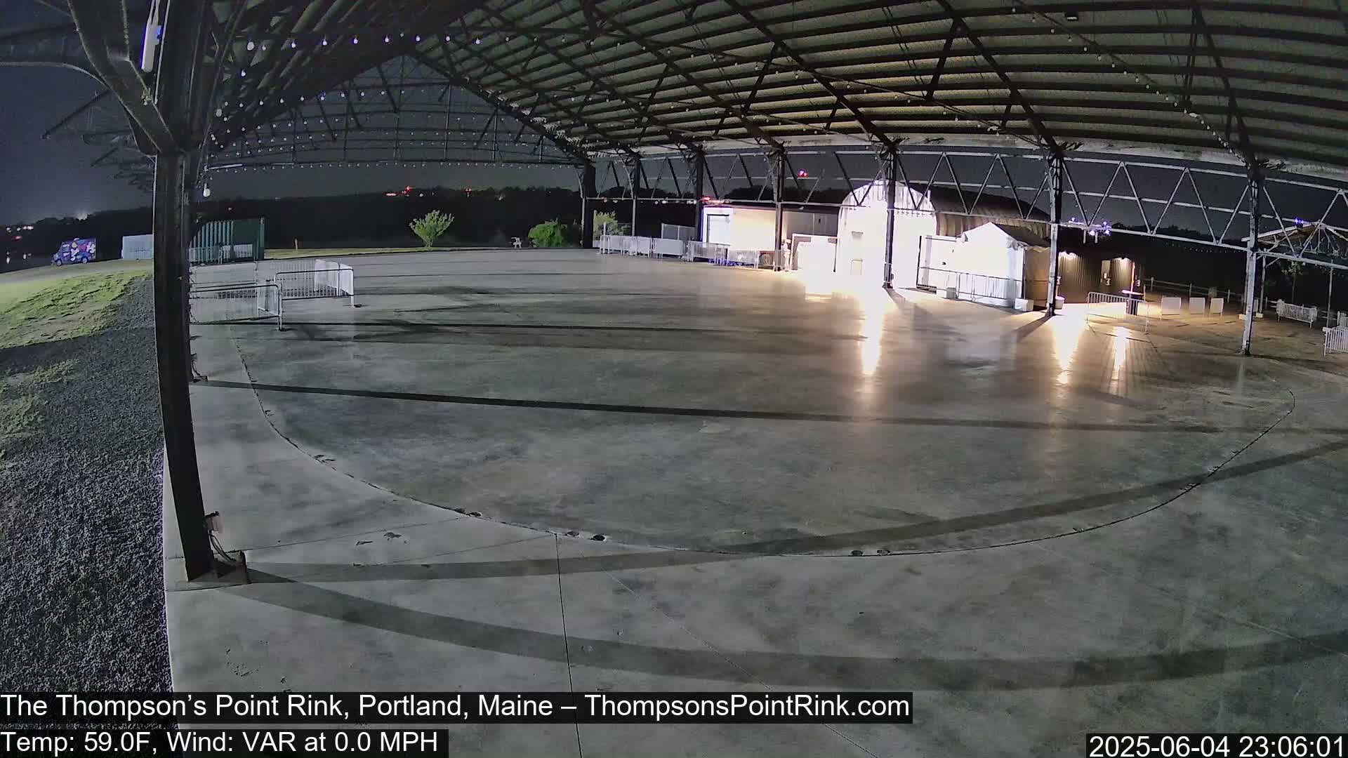 An empty, paved outdoor rink sits under a metal-framed structure at night, with calm weather.