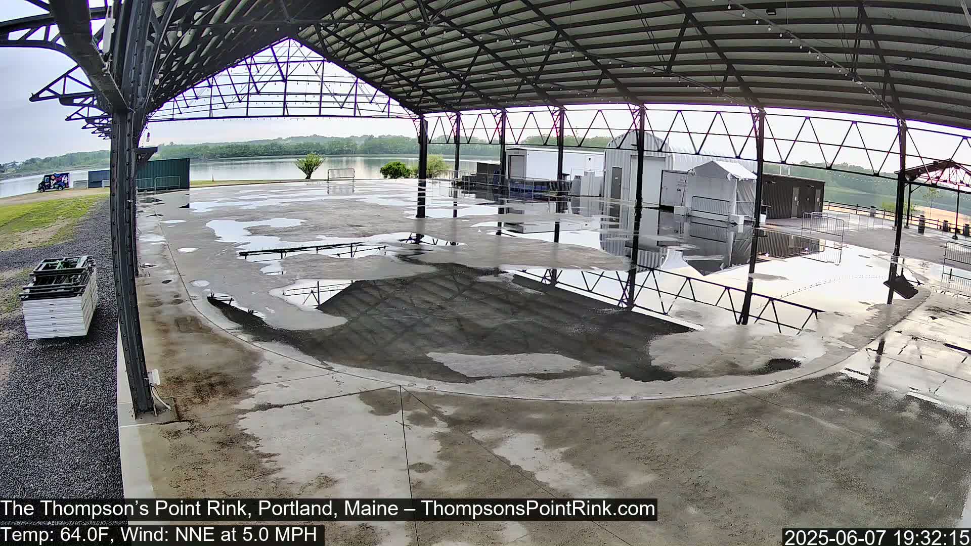 A wet, empty rink under a large metal structure near a body of water on an overcast day.