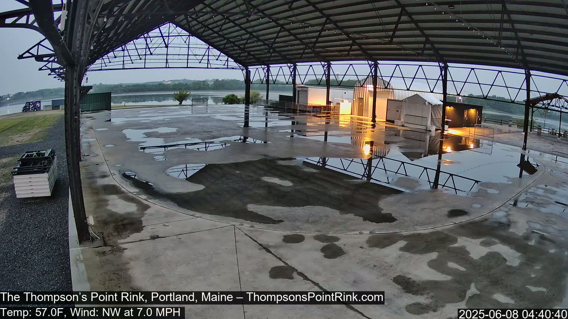 A wet, open-air rink under a metal roof, with a calm body of water visible in the background under an overcast sky.