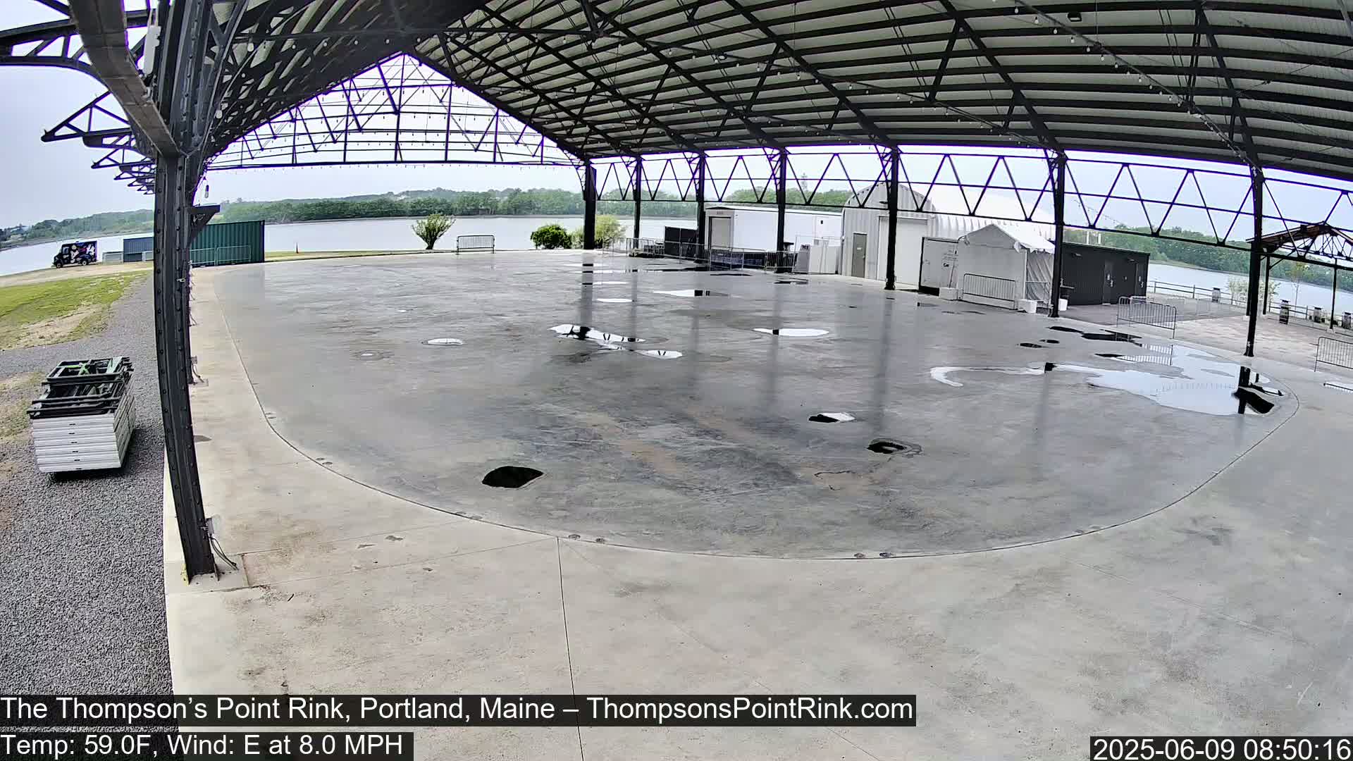 A large, empty, wet concrete rink is under a metal-framed covered structure, with a body of water visible in the background.