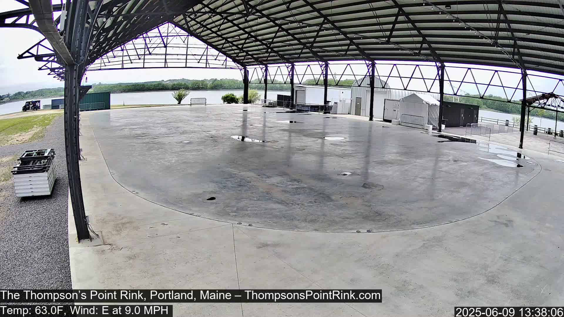 A large, empty, wet concrete rink is situated under a metal-framed structure, overlooking a calm body of water under a mostly clear sky.