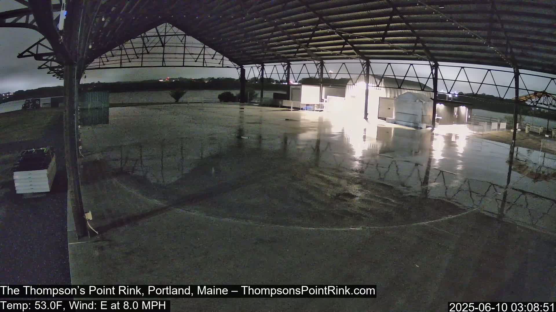 A wet, empty rink sits under a metal roof at night during a light rain with an easterly wind.