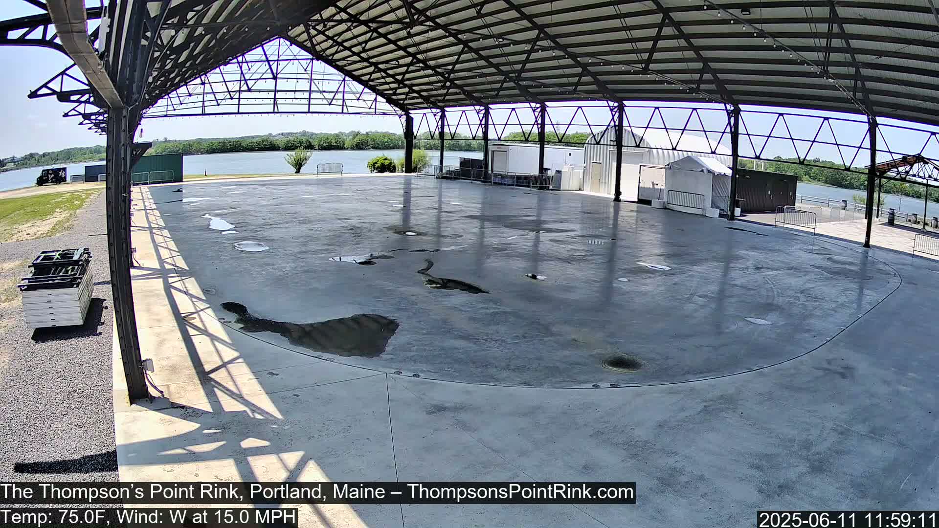 A large, empty, paved rink under a metal-framed shelter on a sunny day with a 15 mph west wind.