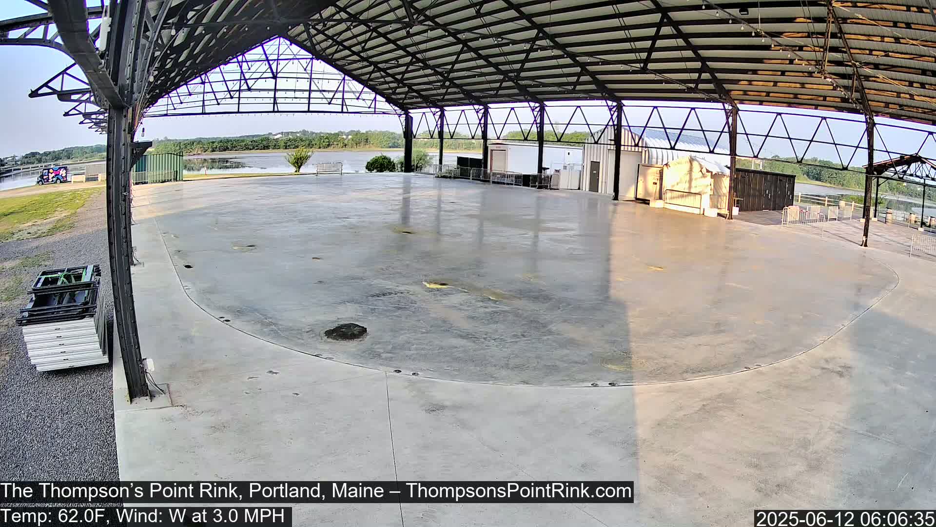 A large, empty, concrete rink sits under a metal-framed shelter on a calm, sunny day.