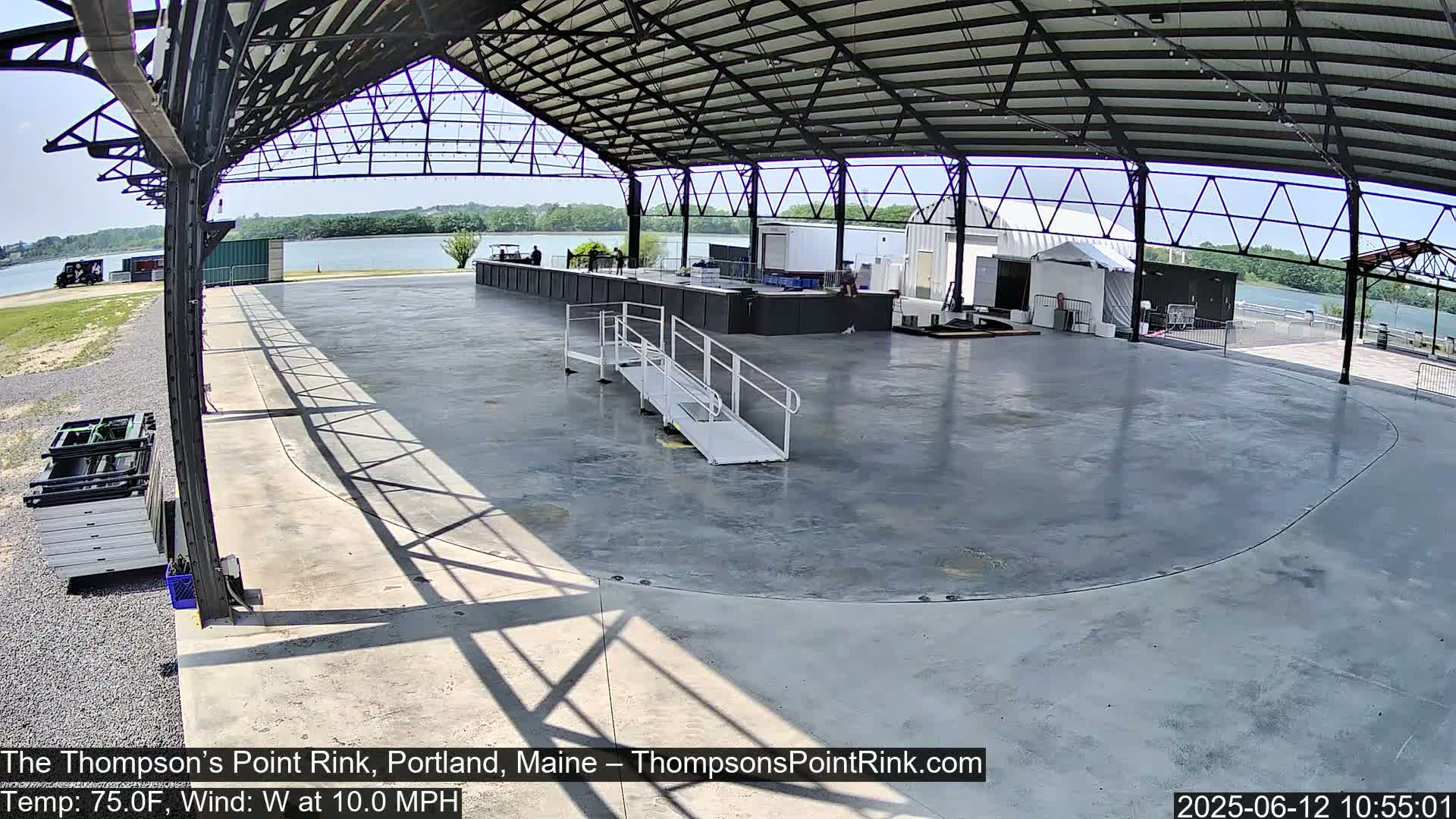 An empty, paved rink under a metal-framed open-air structure on a sunny day with a gentle west wind.