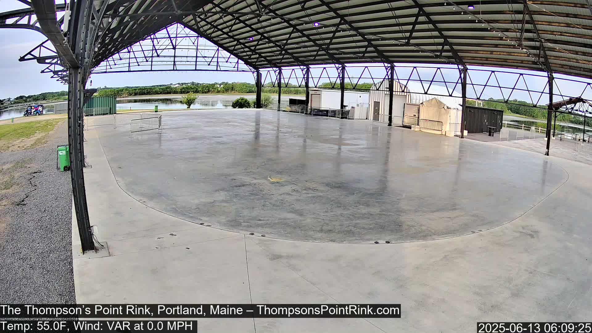 A large, empty concrete rink sits under a metal-framed shelter, next to a calm body of water under an overcast sky.