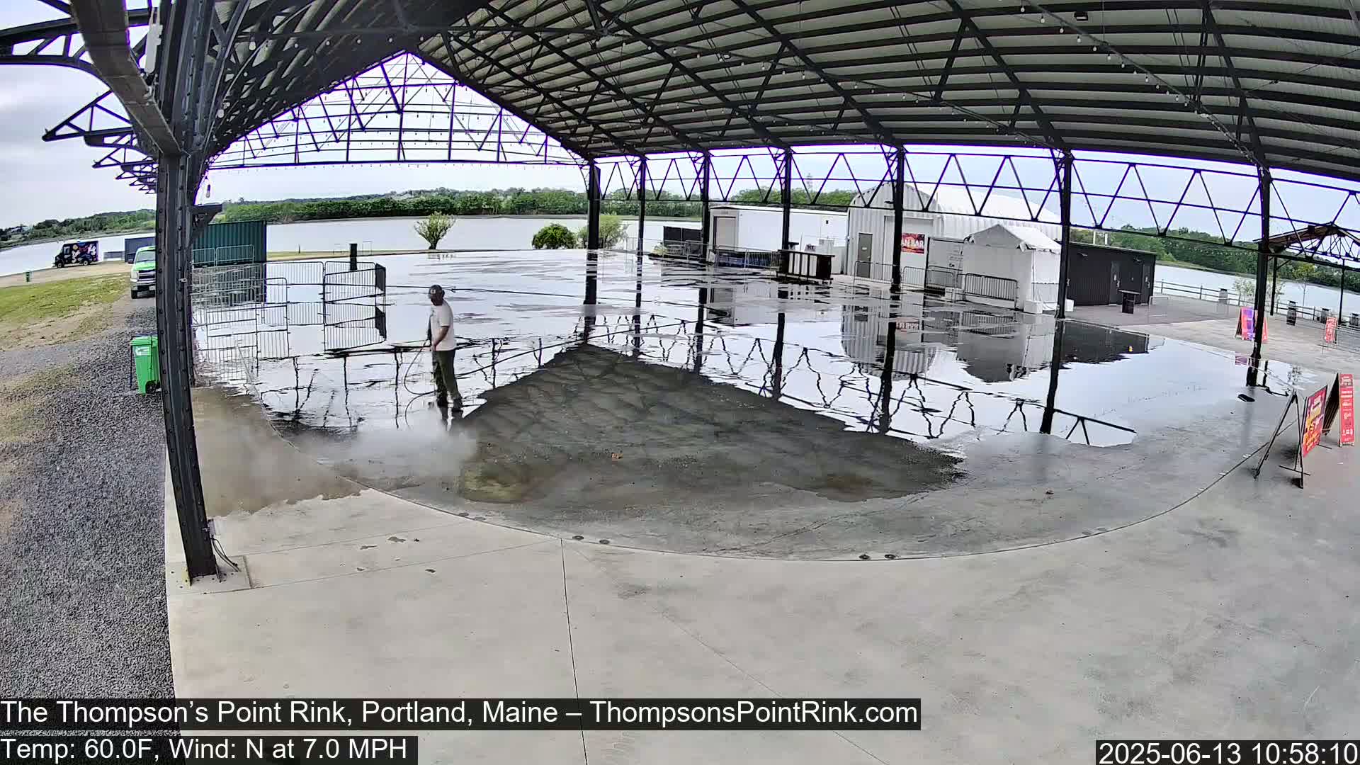 A person is power washing a wet concrete rink under a large metal structure next to a body of water on a partly cloudy day with a north wind.