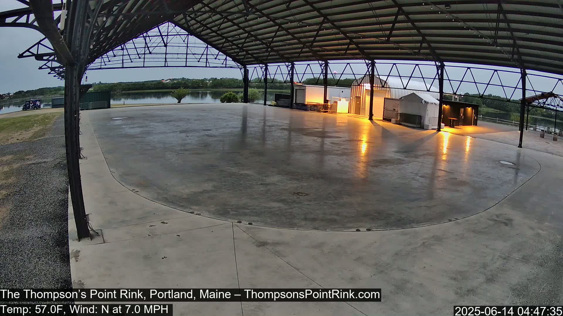 A large, empty, wet concrete rink sits under a metal pavilion at dusk, with a calm body of water visible in the background.