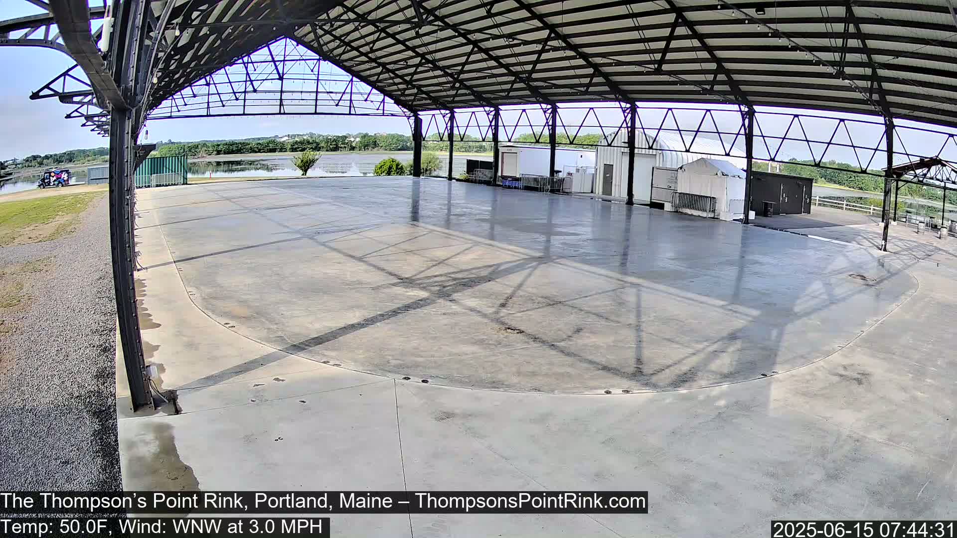 A large, empty, concrete rink sits under a metal-framed shelter on a partly sunny day with a light wind.