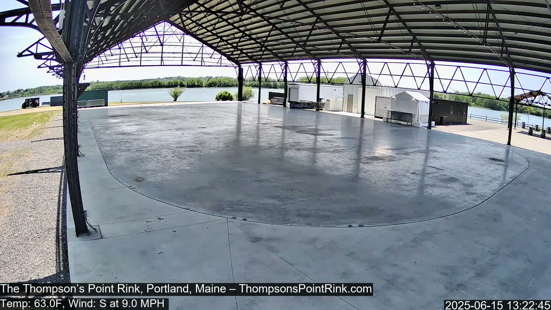 A large, empty, gray concrete rink sits under a metal-framed open-air structure on a sunny day with a calm body of water visible in the background.