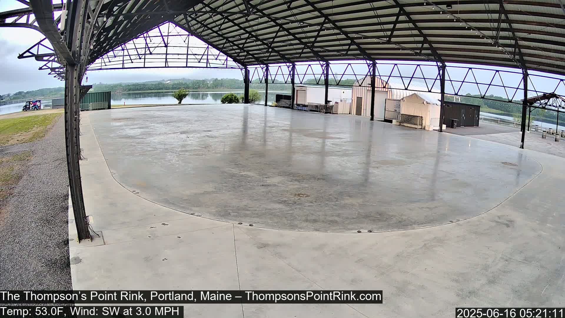 A large, empty, concrete rink sits under a metal-framed structure overlooking a calm body of water on an overcast day.