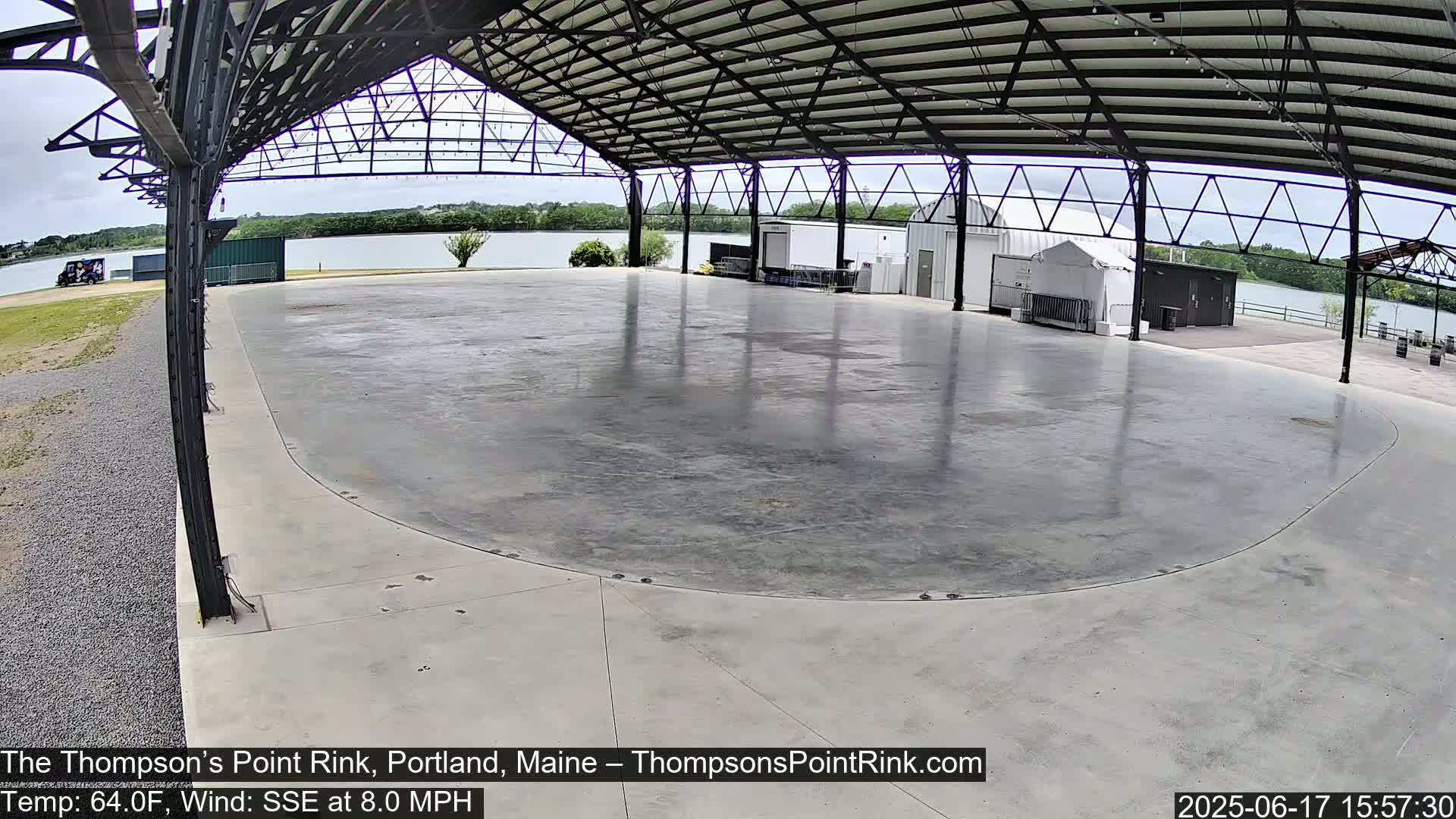 A large, empty, concrete rink sits under a metal-framed shelter, overlooking a calm body of water on a partly cloudy day with a SSE wind at 8 mph.