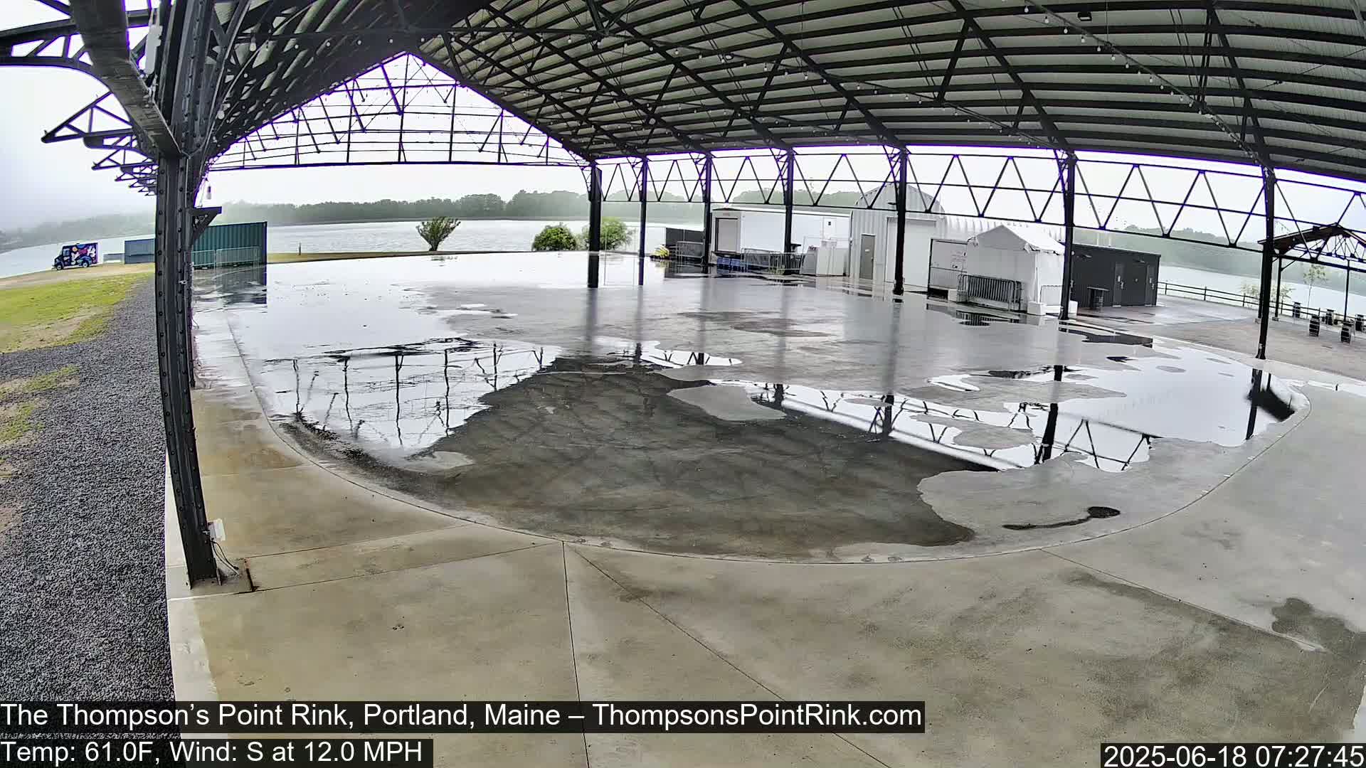 A wet, open-air rink under a metal roof overlooks a calm body of water during a rainy day with a south wind.