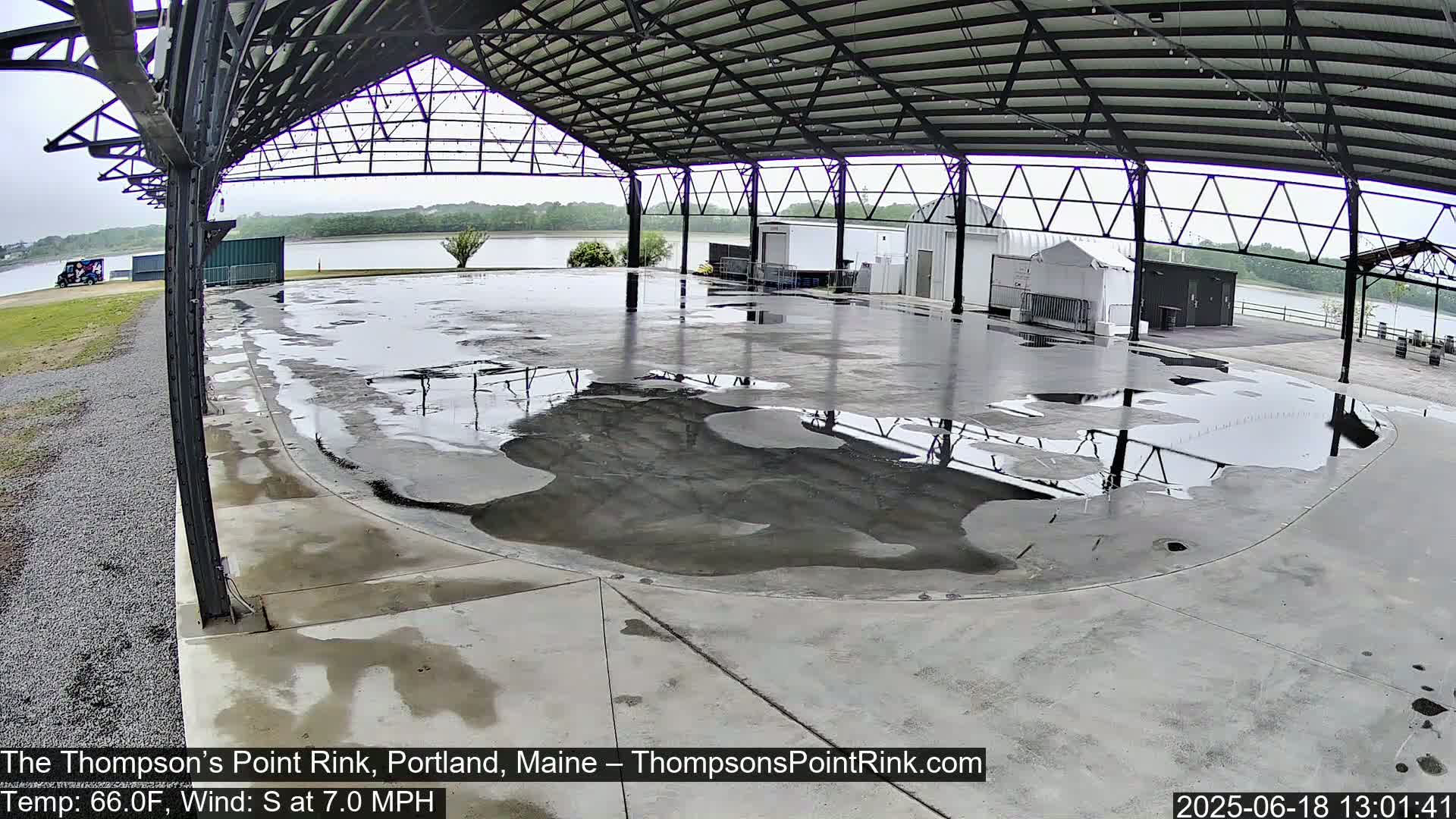 A wet, open-air concrete rink sits under a metal framework, with a calm body of water visible in the background under an overcast sky.