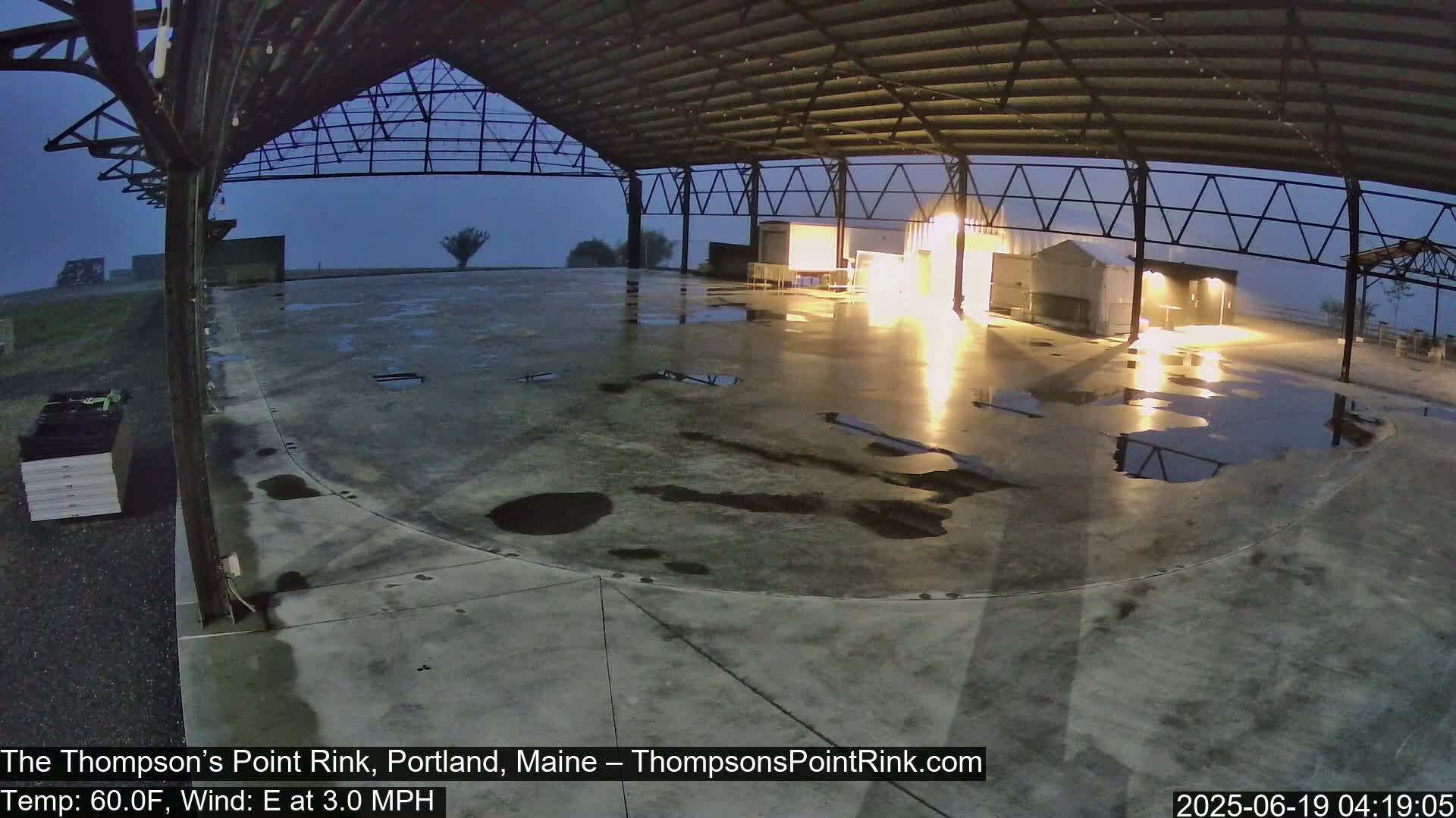 A wet, empty paved area under a large metal structure at night, with a light mist or fog.