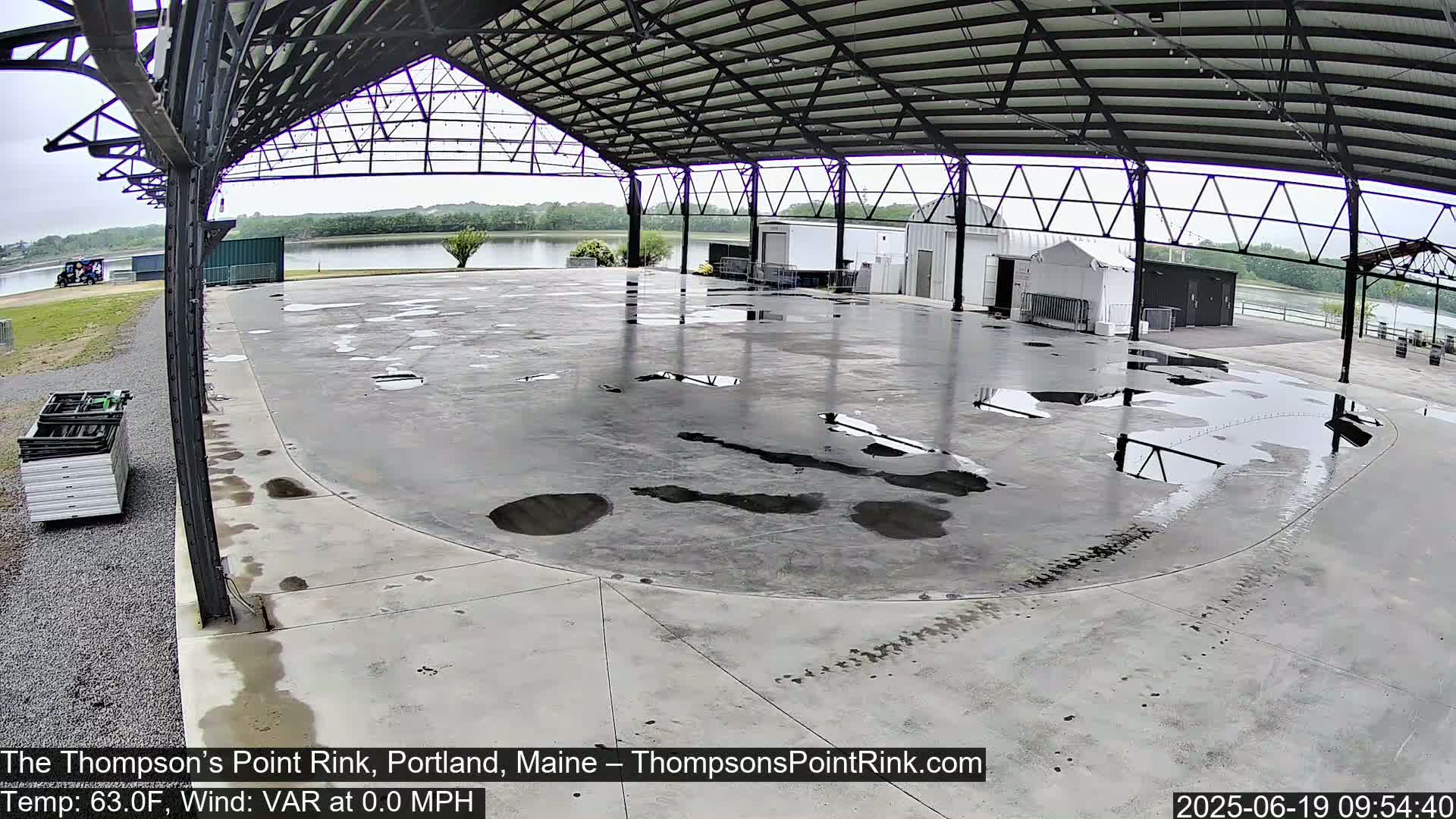 A wet, empty concrete rink sits under a metal-framed shelter, overlooking a calm body of water on an overcast day.