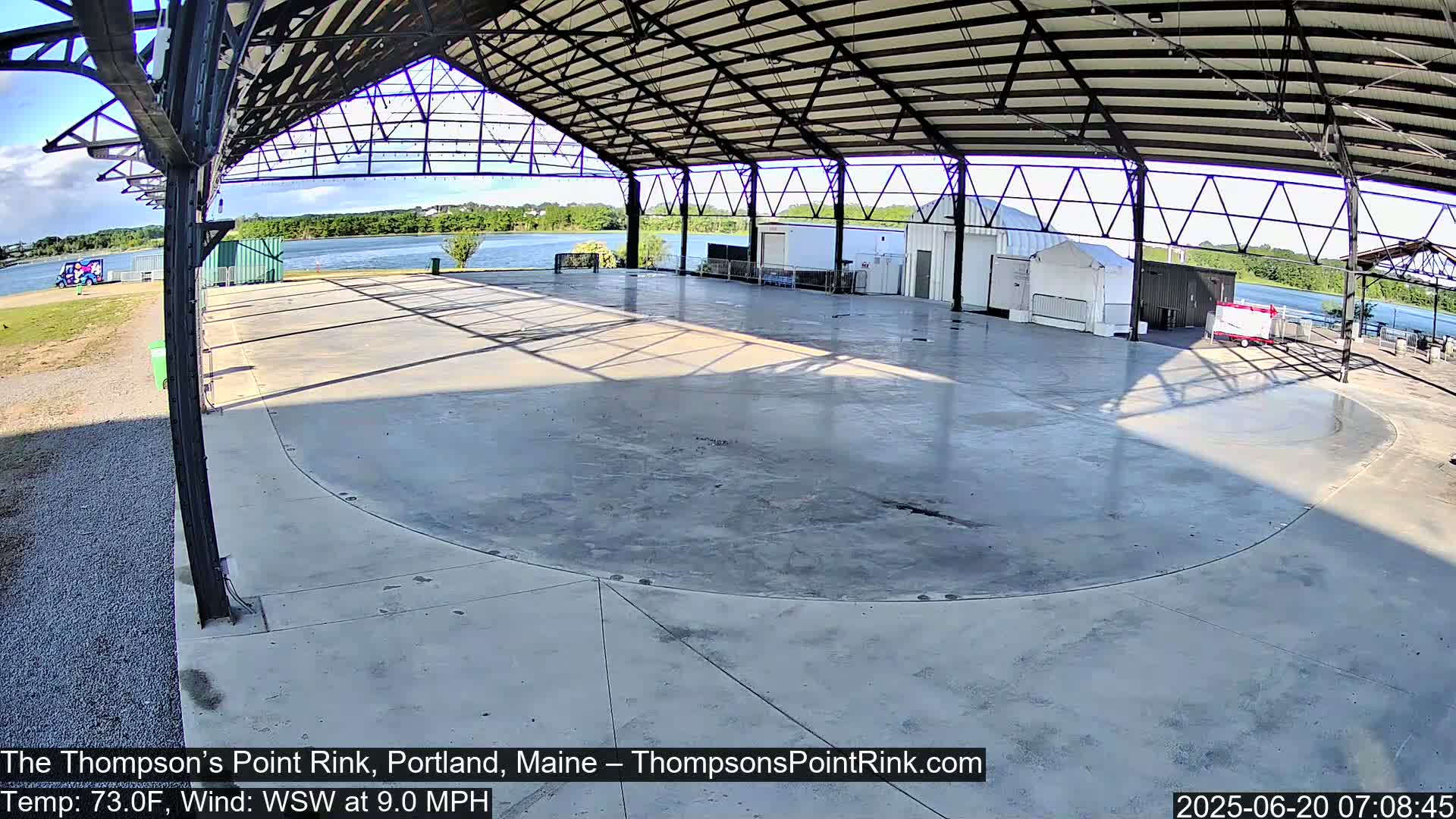 A large empty paved rink sits under a metal awning beside a calm body of water on a partly sunny day with a light wind.