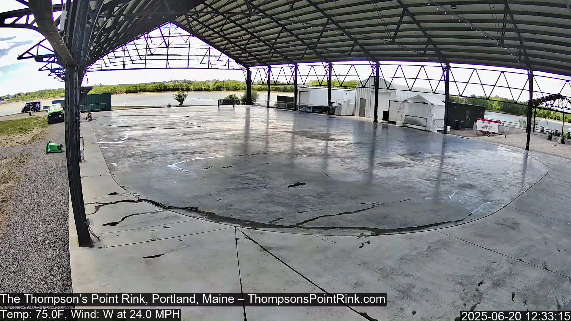 A large, cracked concrete rink sits under a metal awning, overlooking a calm river under a partly cloudy sky with a 24 mph west wind.