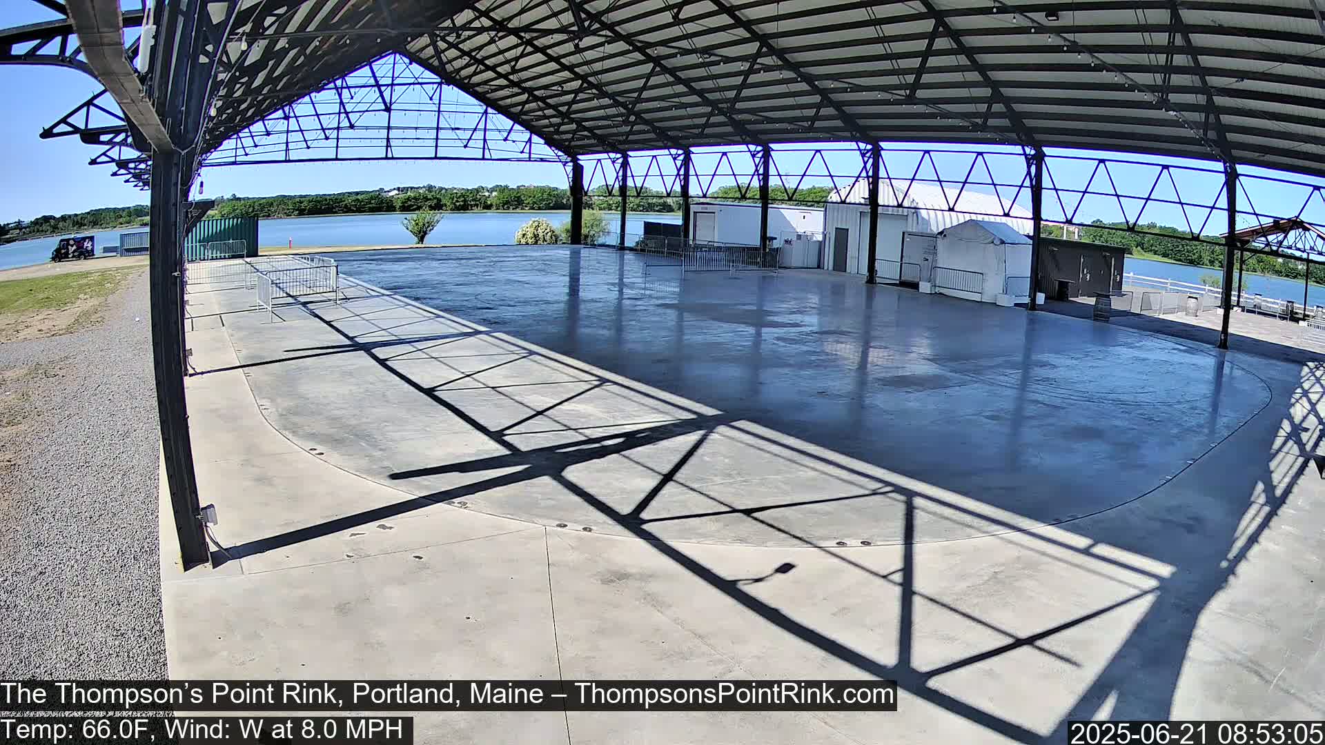An empty, paved rink under a metal-framed shelter overlooks a calm lake on a sunny day.