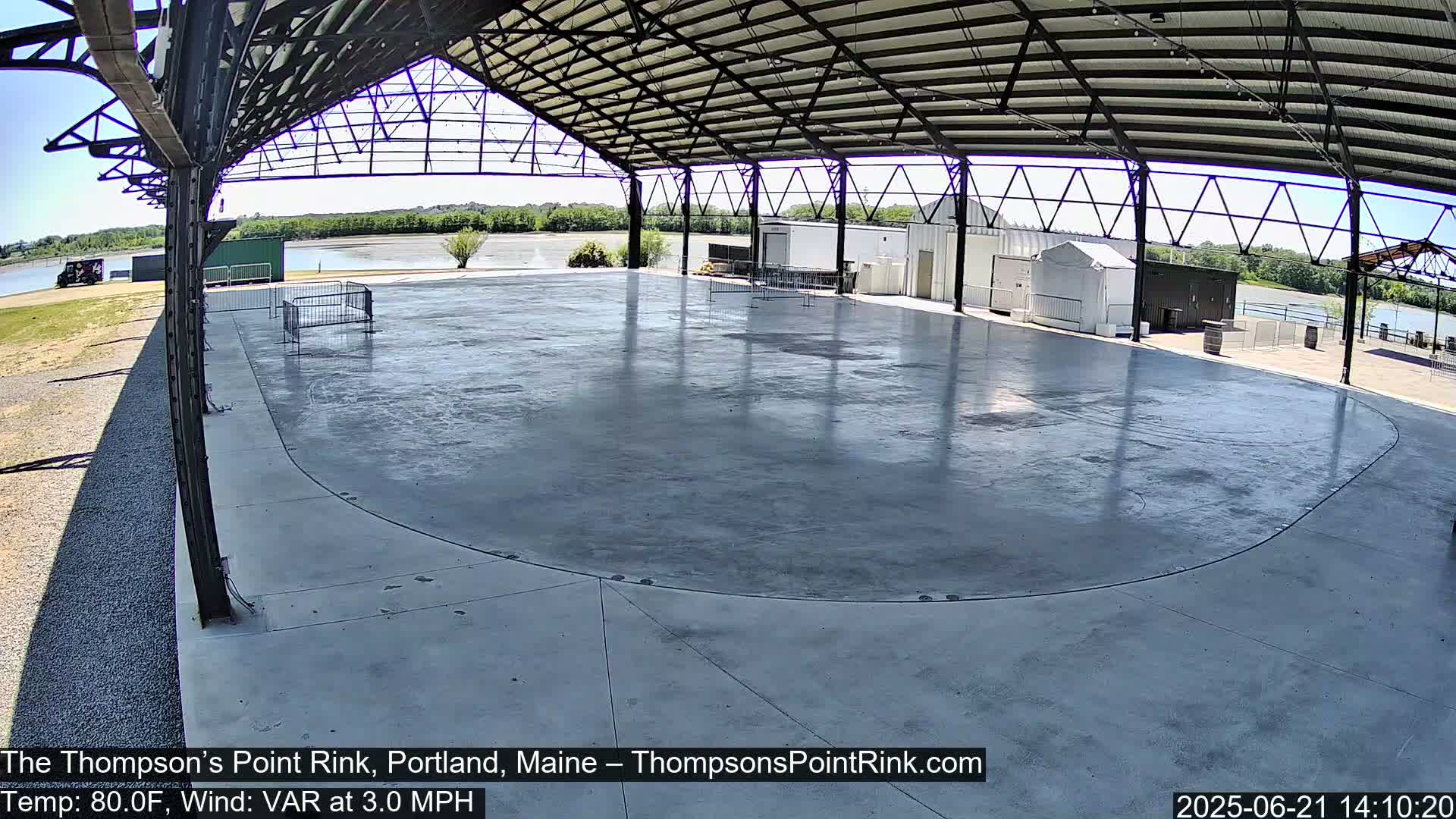 An empty, curved, outdoor concrete rink sits under a metal roof on a sunny day with a light wind.