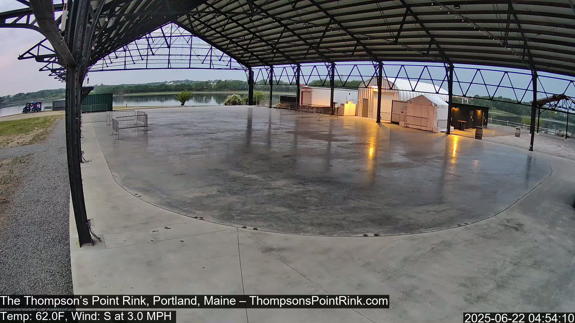 An empty, wet concrete rink sits under a metal roof structure, overlooking a calm body of water under an overcast sky.