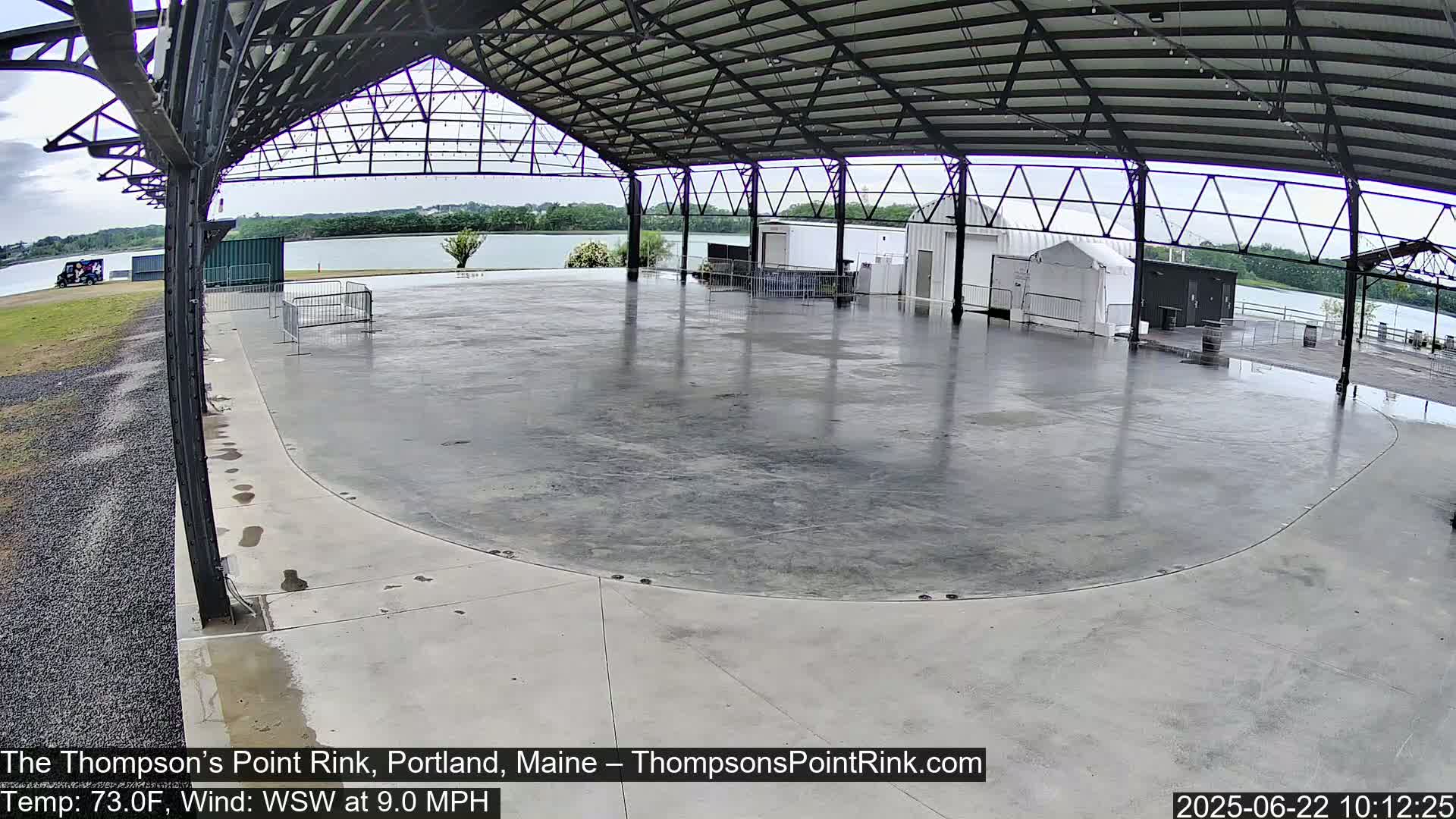 A wet, empty concrete rink sits under a metal roofed structure overlooking a calm body of water on a partly cloudy day.