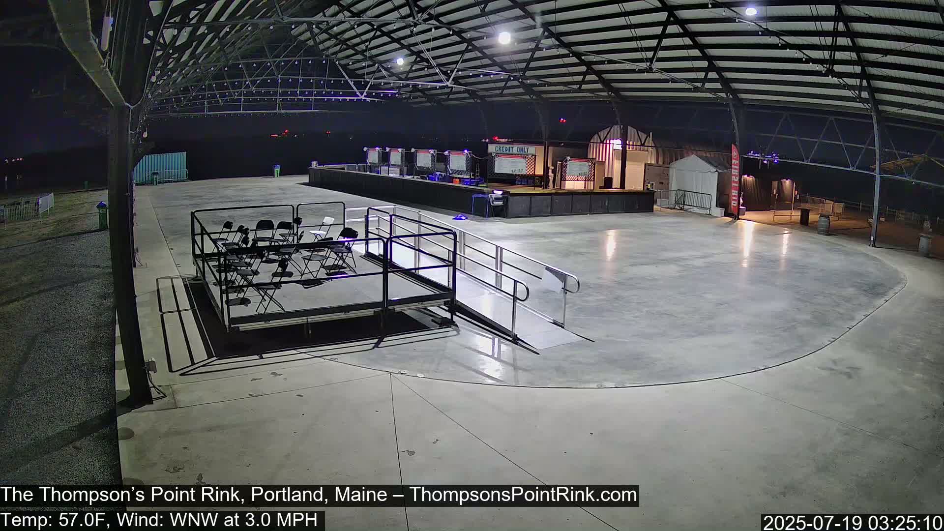 An empty, paved rink under a metal-framed building at night, illuminated by artificial lights, with a few chairs set up on a small stage and a ramp leading to it.