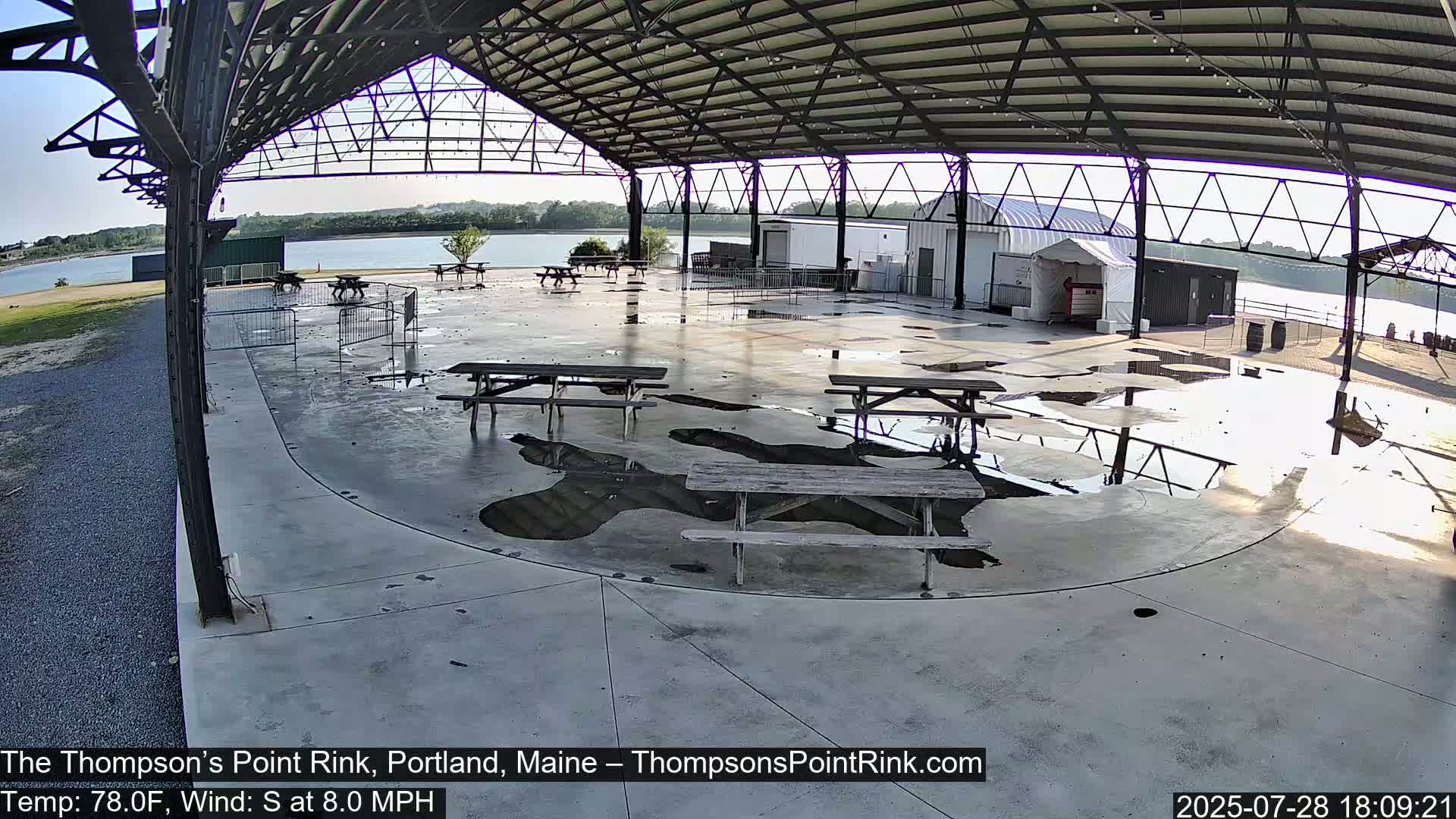 An open-air pavilion with several picnic tables sits near a body of water under a partly cloudy sky, with puddles on the ground indicating recent rain.