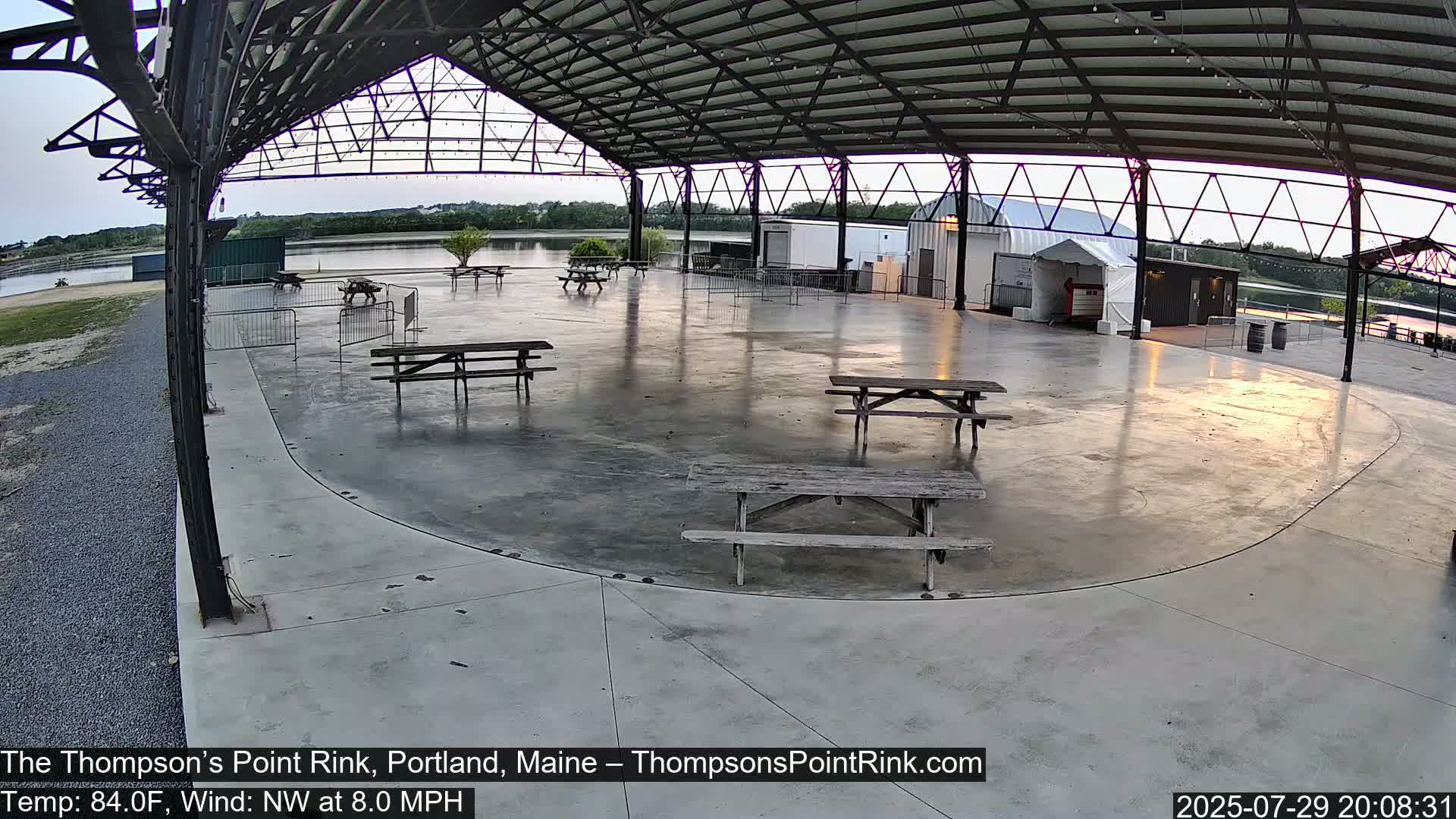 An open-air pavilion with several picnic tables sits near a body of water under a partly cloudy sky, with puddles on the ground indicating recent rain.