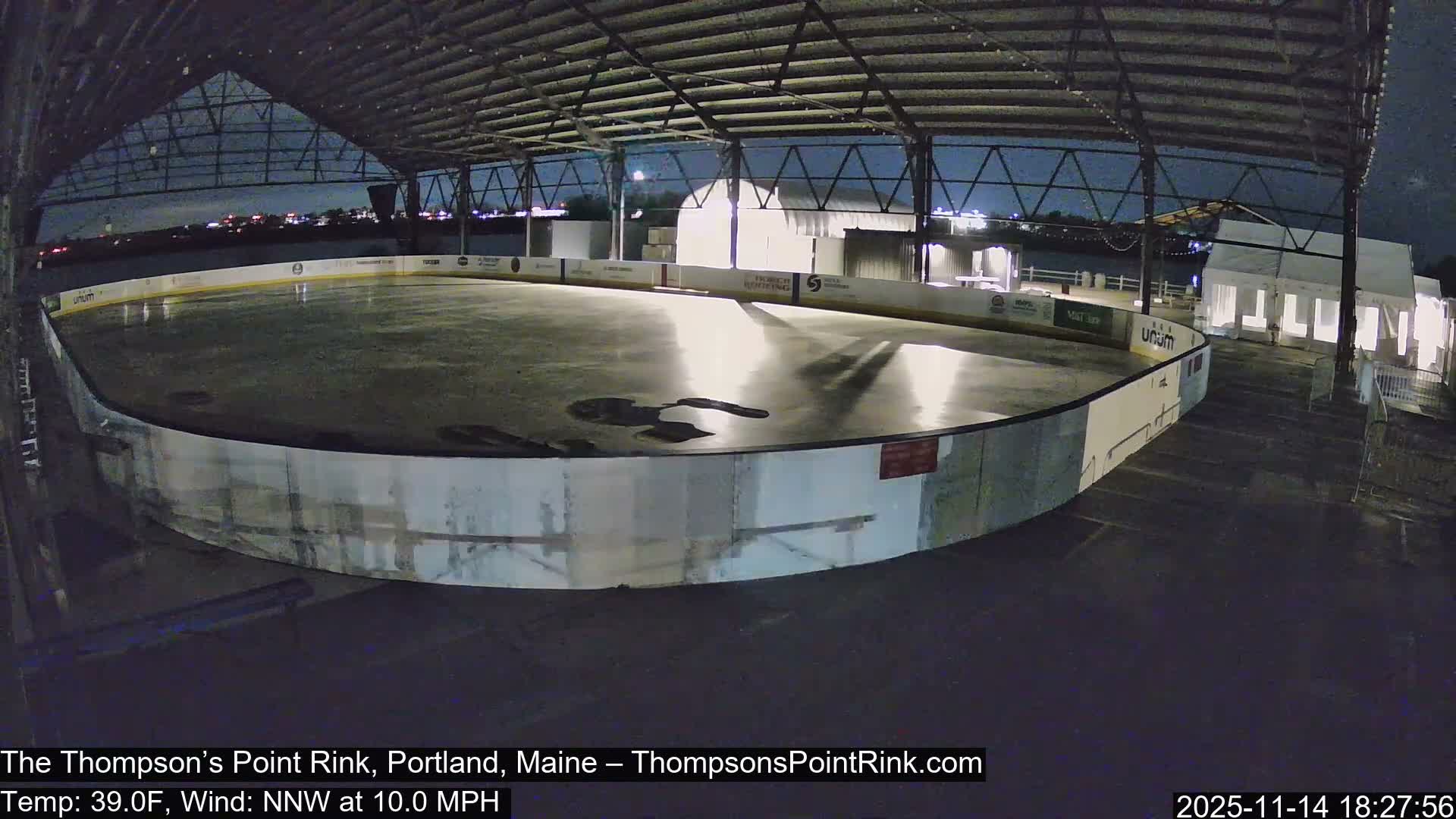 An empty, partially melted outdoor ice rink with puddles is visible at night under an open-sided roof structure, with a temperature of 39.0F and a 10.0 MPH NNW wind.