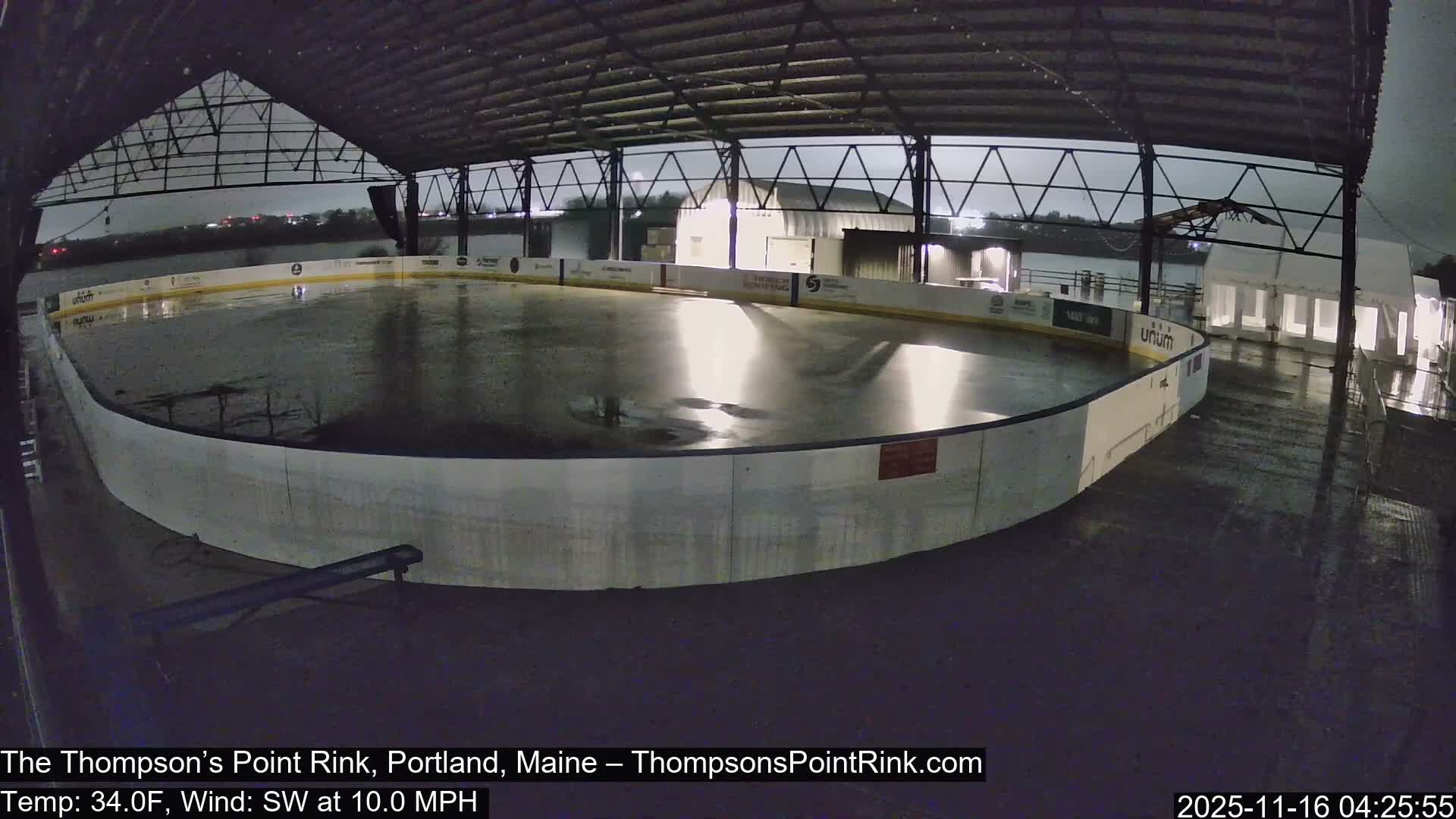 An empty, covered ice rink appears wet and reflective under a dark, overcast sky, indicating rainy conditions with a temperature of 34.0F and a SW wind at 10.0 MPH.
