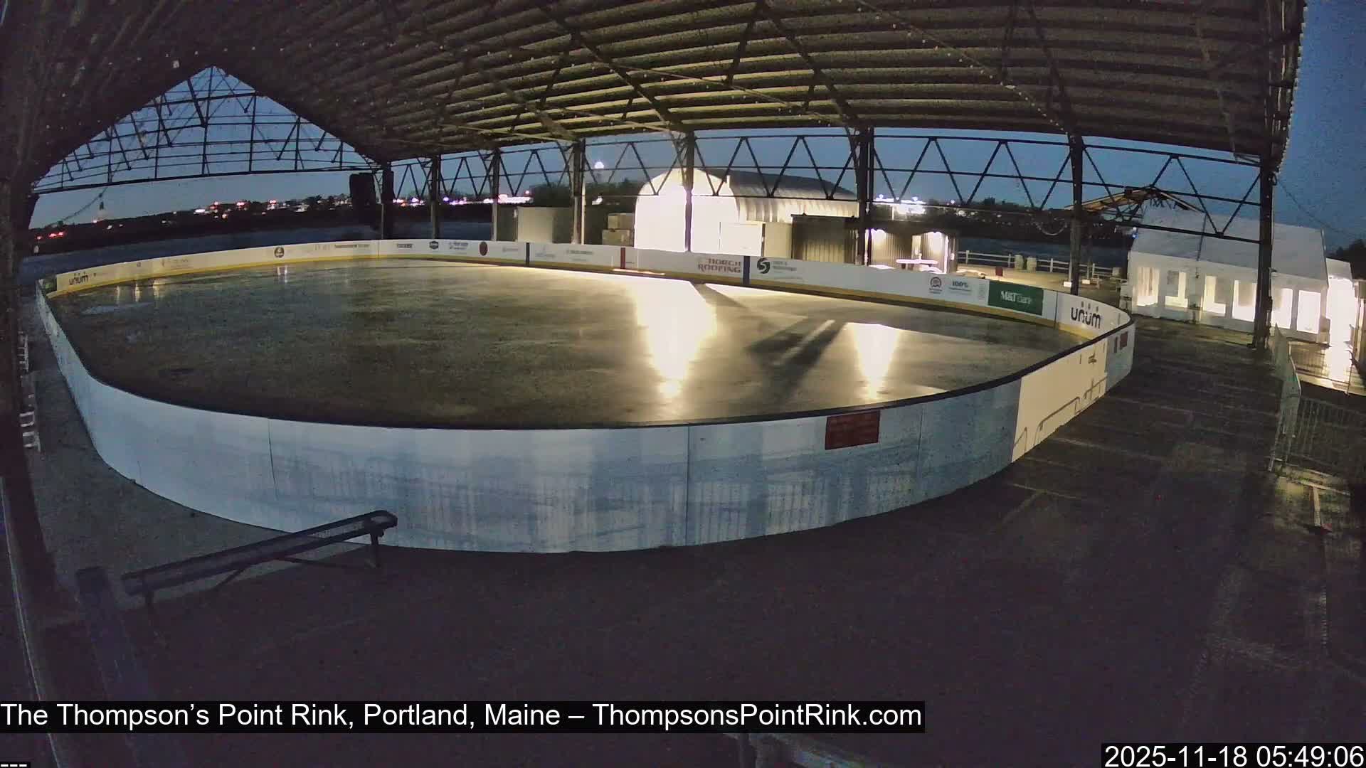 An empty, covered ice rink appears wet and reflective under a dark, overcast sky, indicating rainy conditions with a temperature of 34.0F and a SW wind at 10.0 MPH.