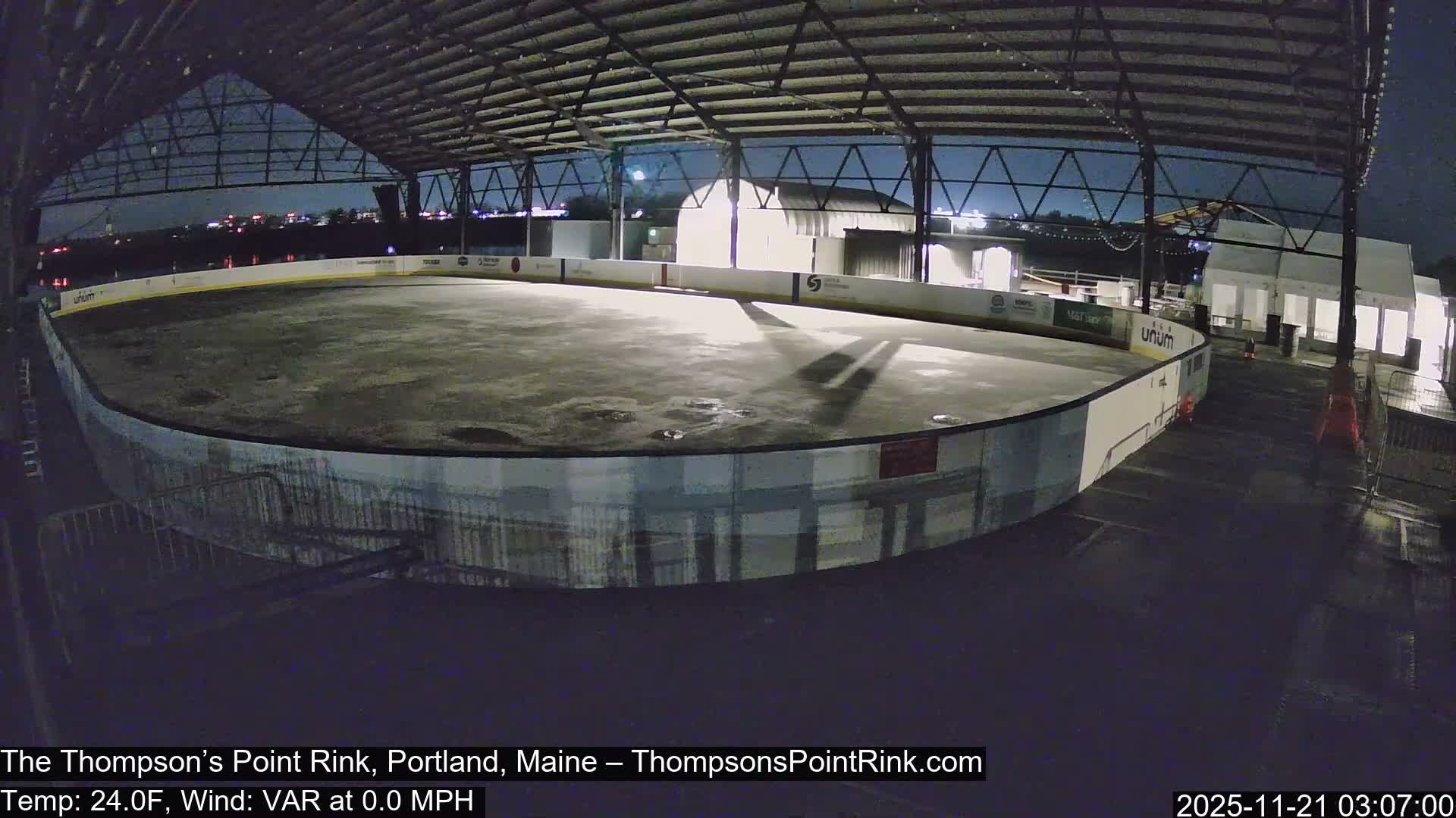 An empty, covered outdoor ice rink, its base surface appearing wet with puddles and dirt, is illuminated by artificial lights on a clear, dark night, with distant city lights visible beyond the open structure.