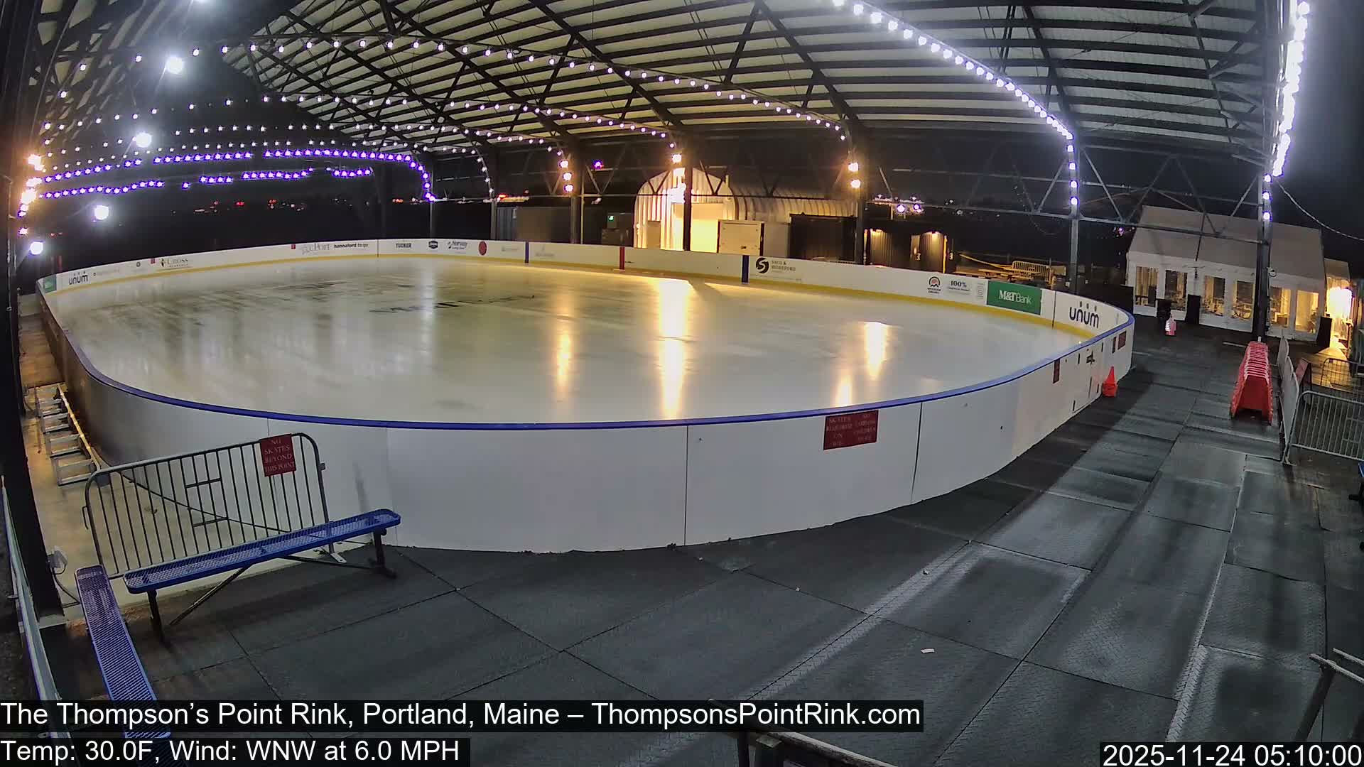 A brightly lit, empty outdoor ice rink, covered by a large structure adorned with string lights, is visible at night with a temperature of 30.0F and a WNW wind at 6.0 MPH.
