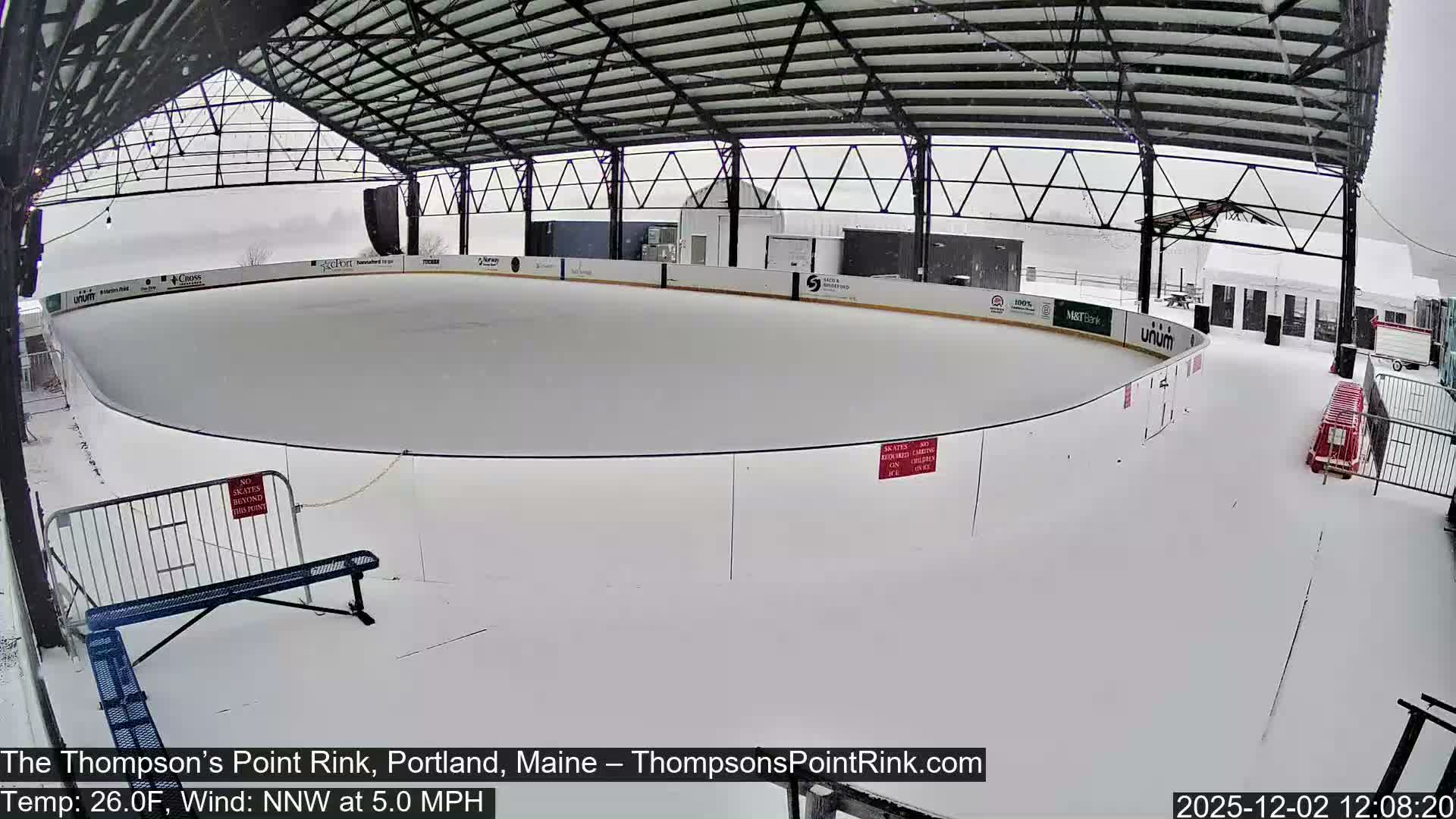 An outdoor ice rink under an open-sided roof structure is entirely blanketed in snow under a grey, overcast sky with active snowfall.