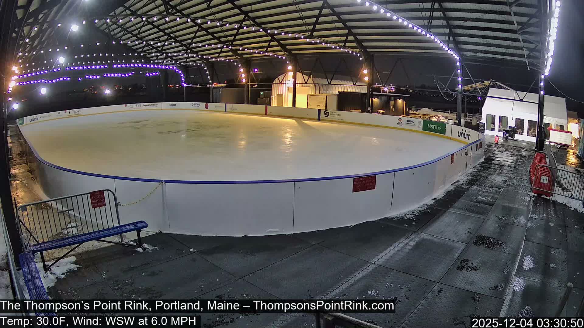 An empty outdoor ice skating rink is brightly illuminated under a large, open-sided shelter at night, with patches of snow visible on the ground around it.
