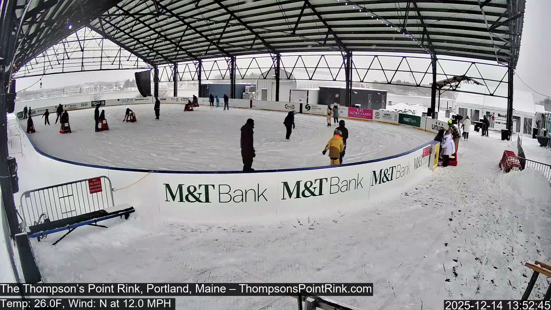 An empty outdoor ice skating rink is brightly illuminated under a large, open-sided shelter at night, with patches of snow visible on the ground around it.