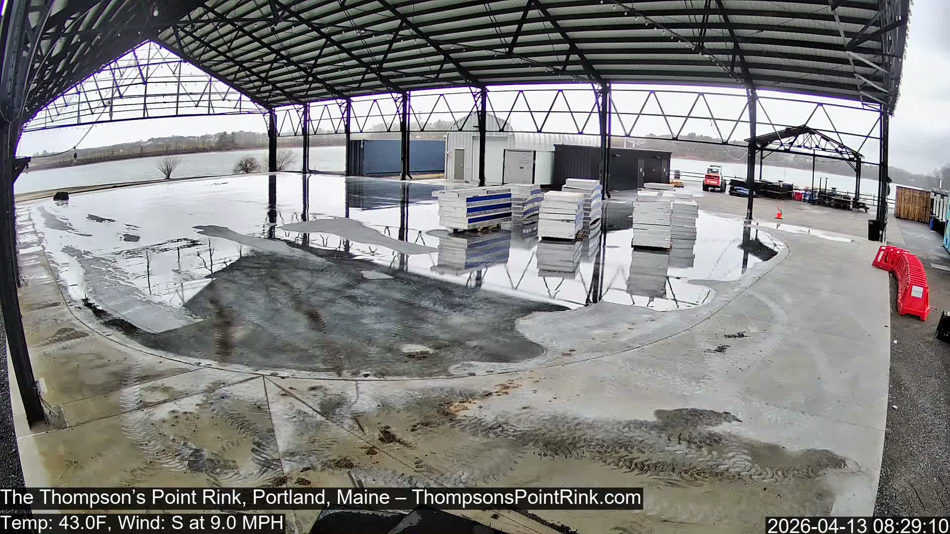 An empty outdoor ice skating rink is brightly illuminated under a large, open-sided shelter at night, with patches of snow visible on the ground around it.