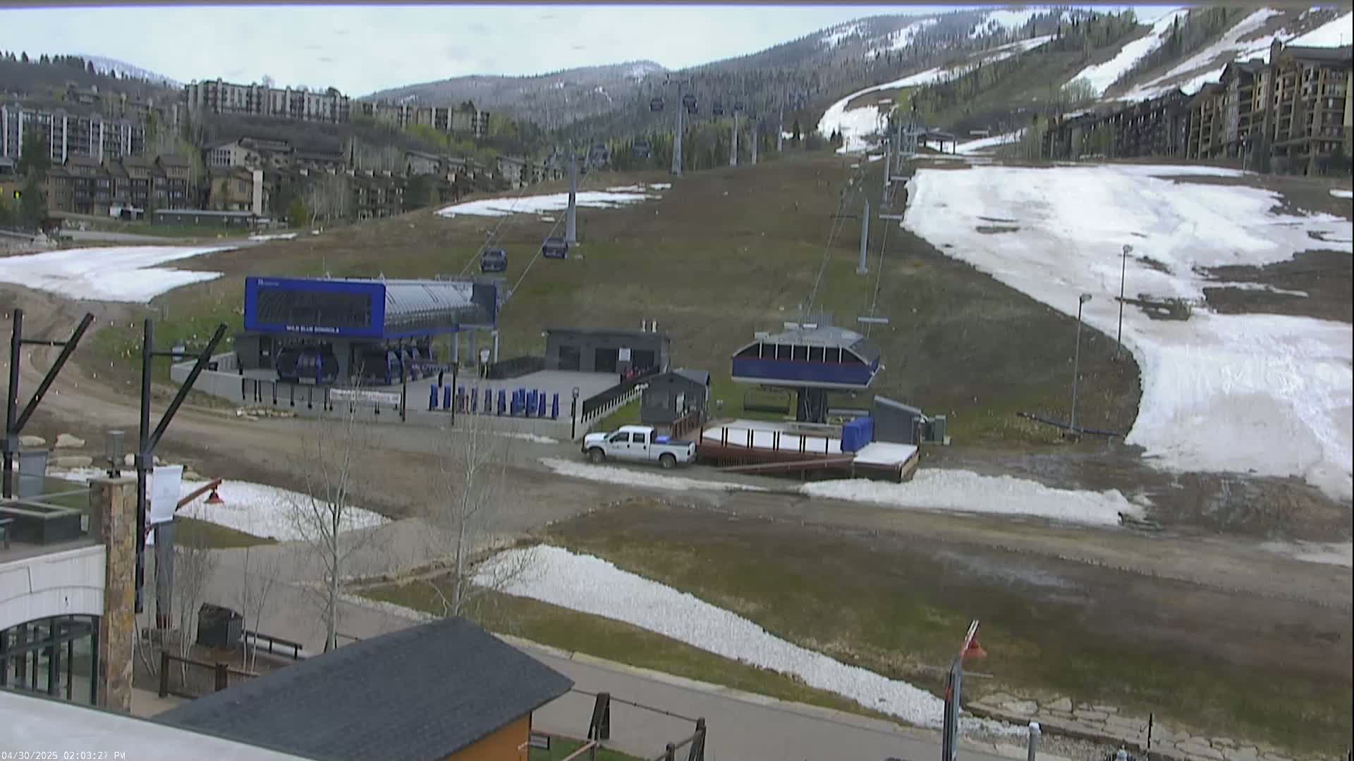 A mostly clear day shows a ski resort with melting snow patches, several chairlifts, and buildings in the background.