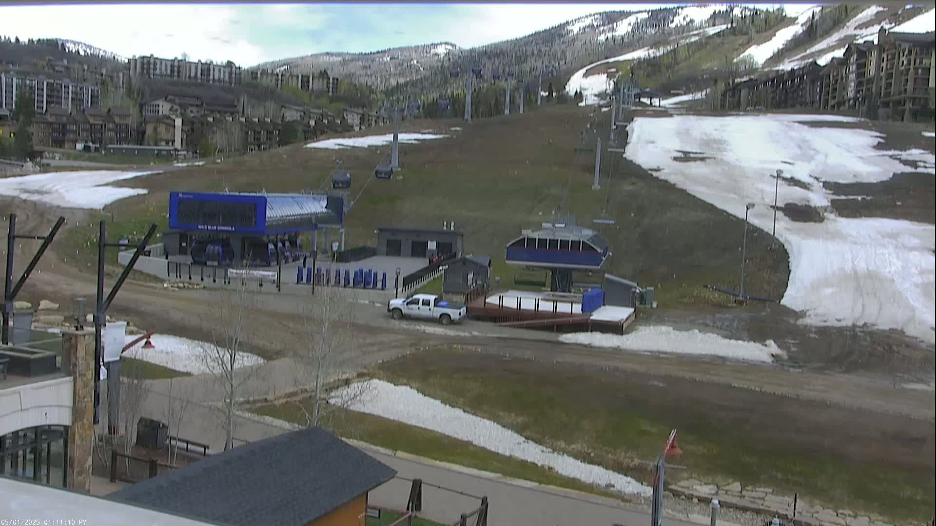 A mostly clear day shows a ski lift base area with patchy snow, a white pickup truck, and buildings in the background.