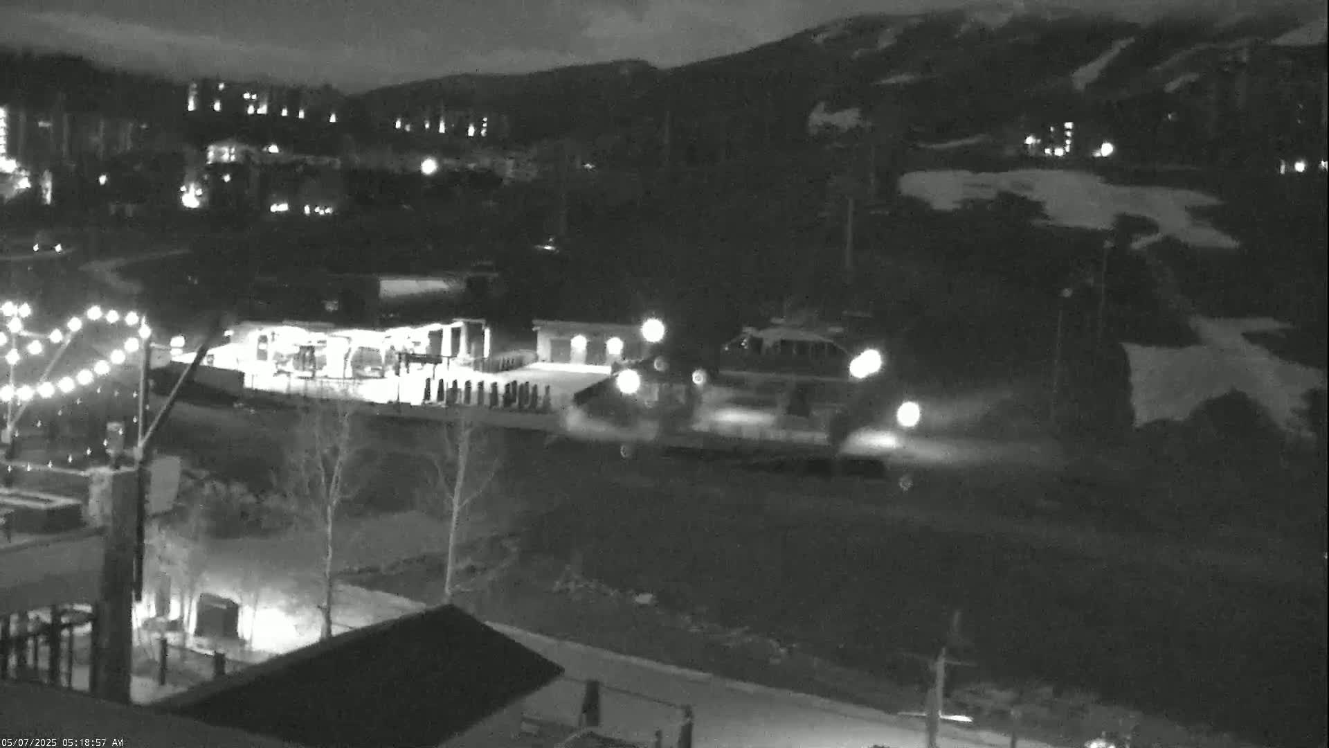 A nighttime, snow-covered scene shows vehicles with headlights illuminated near a building and string lights, with a mountainside in the background.