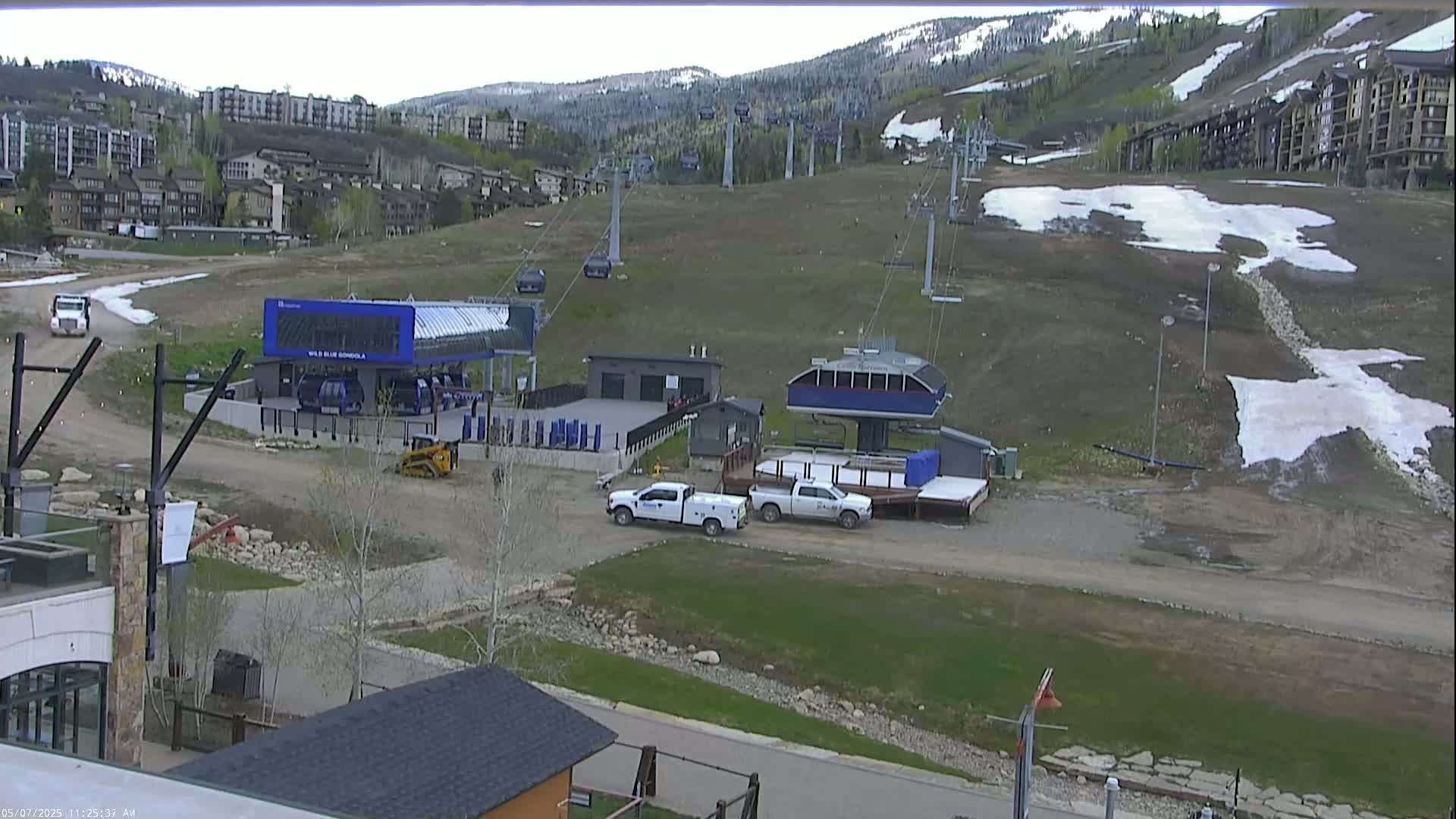 A mostly clear day shows a ski resort with two chairlifts, some buildings, and a few vehicles in a parking area, with patches of remaining snow on the slopes and surrounding ground.
