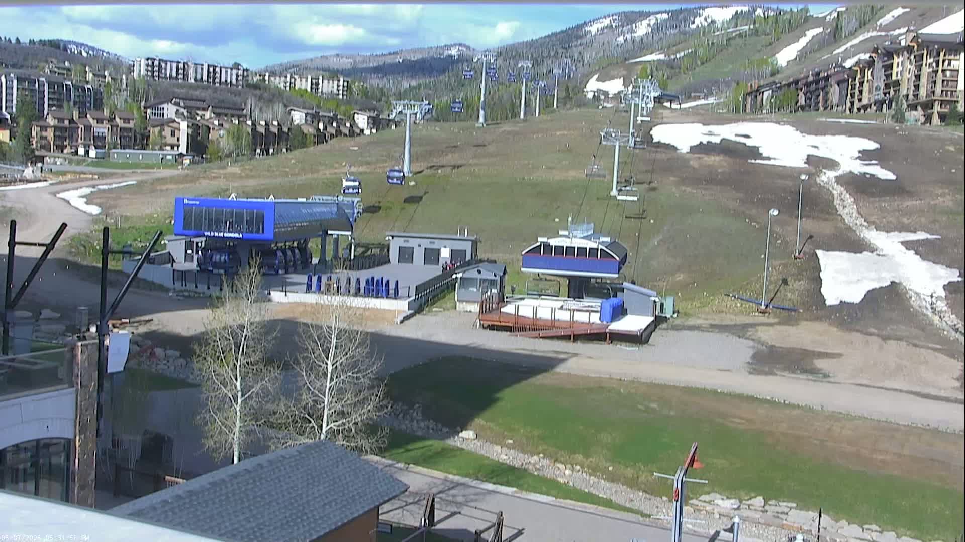 A mostly sunny day reveals a ski resort base area with several chairlifts, buildings, and patches of remaining snow.