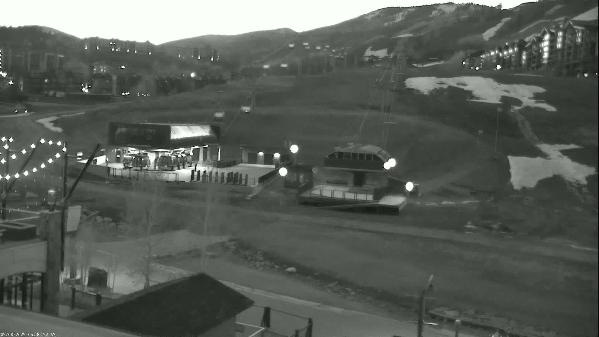 A nighttime, grayscale image shows a ski resort with a chairlift ascending a snow-dusted hill toward buildings lit with interior lights, under a dark, clear sky.