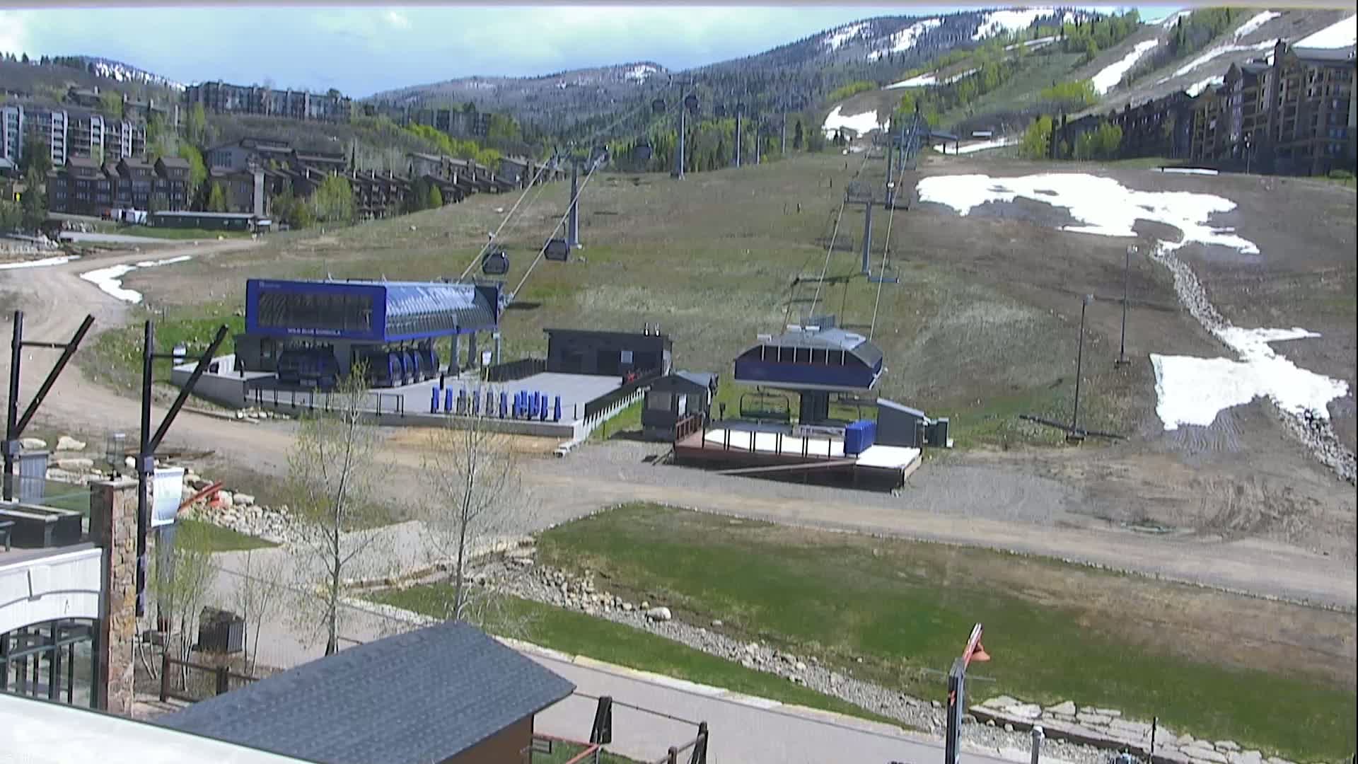 A mostly clear day shows two ski lift stations and a mostly barren ski slope with patches of remaining snow, backed by a mountainside with buildings and trees.