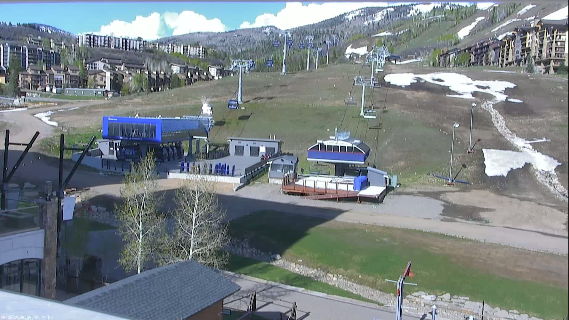 A mostly sunny day reveals a ski resort with several chairlifts, base buildings, and patches of melting snow on the surrounding grounds.
